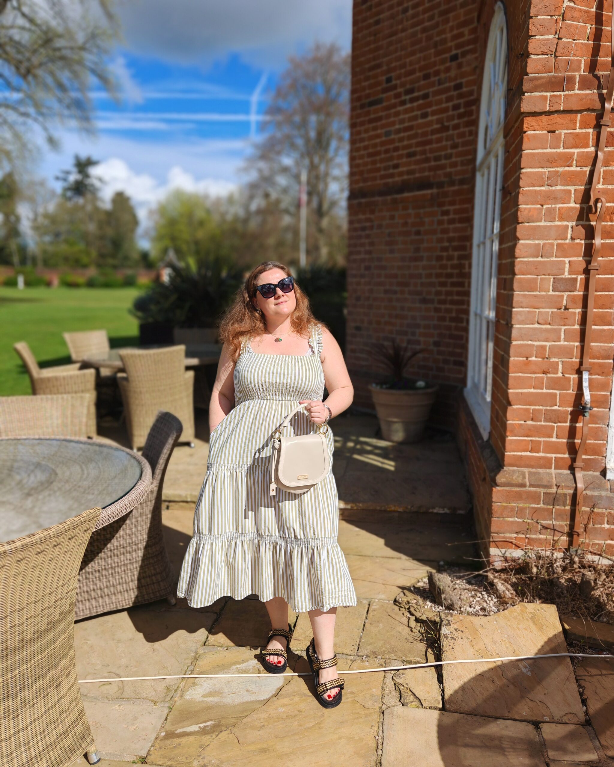 White woman posing near a country house in a stripped green and white mid-dress. She is holding a white leather bag with sunglasses. White woman posing near a country house in a stripped green and white mid-dress. She is holding a white leather bag with sunglasses.
