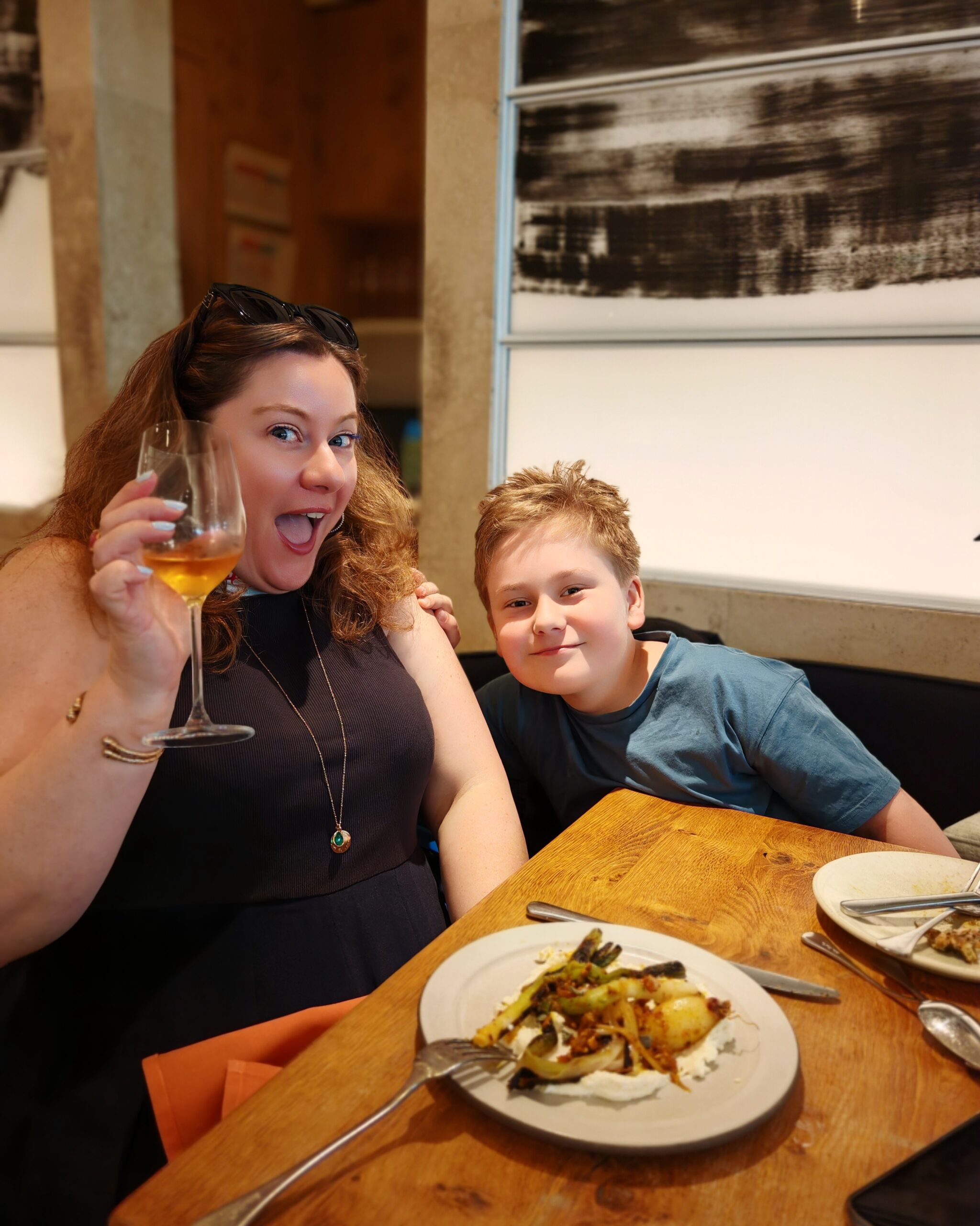 White woman smiling and holding a glass of white whine with a little white boy next to hear. at a restaurant with a plate full of food next to them White woman smiling and holding a glass of white whine with a little white boy next to hear. at a restaurant with a plate full of food next to them