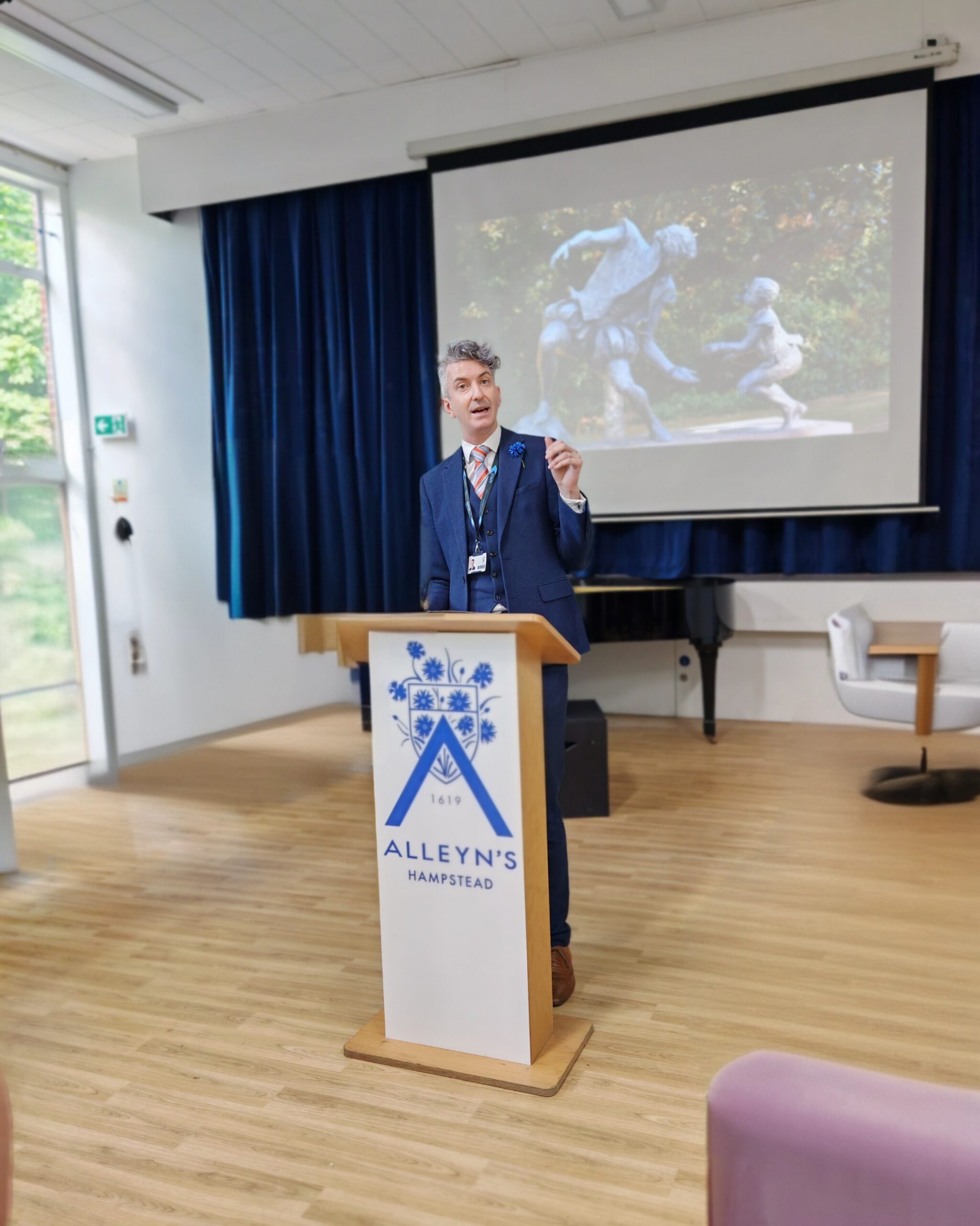 Aidan Sproat-Clements, founding Head of Alleyn's Hampstead giving a speech in the secondary school in North London with the school's group logo in front. Aidan Sproat-Clements, founding Head of Alleyn's Hampstead giving a speech in the secondary school in North London with the school's group logo in front.