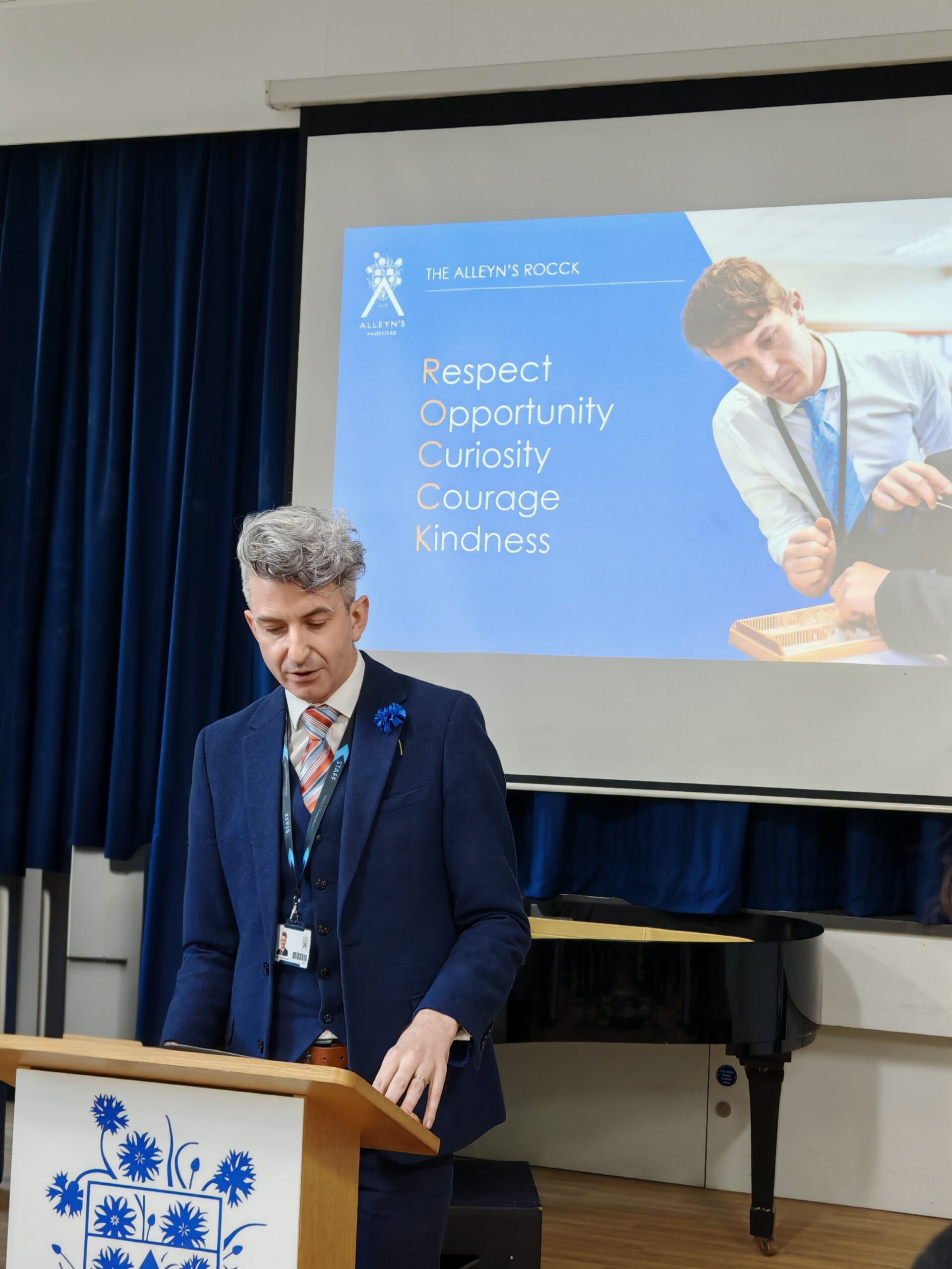 Aidan Sproat-Clements, founding Head of Alleyn's Hampstead giving a speech in the secondary school in North London with the school's group logo in front. Aidan Sproat-Clements, founding Head of Alleyn's Hampstead giving a speech in the secondary school in North London with the school's group logo in front.
