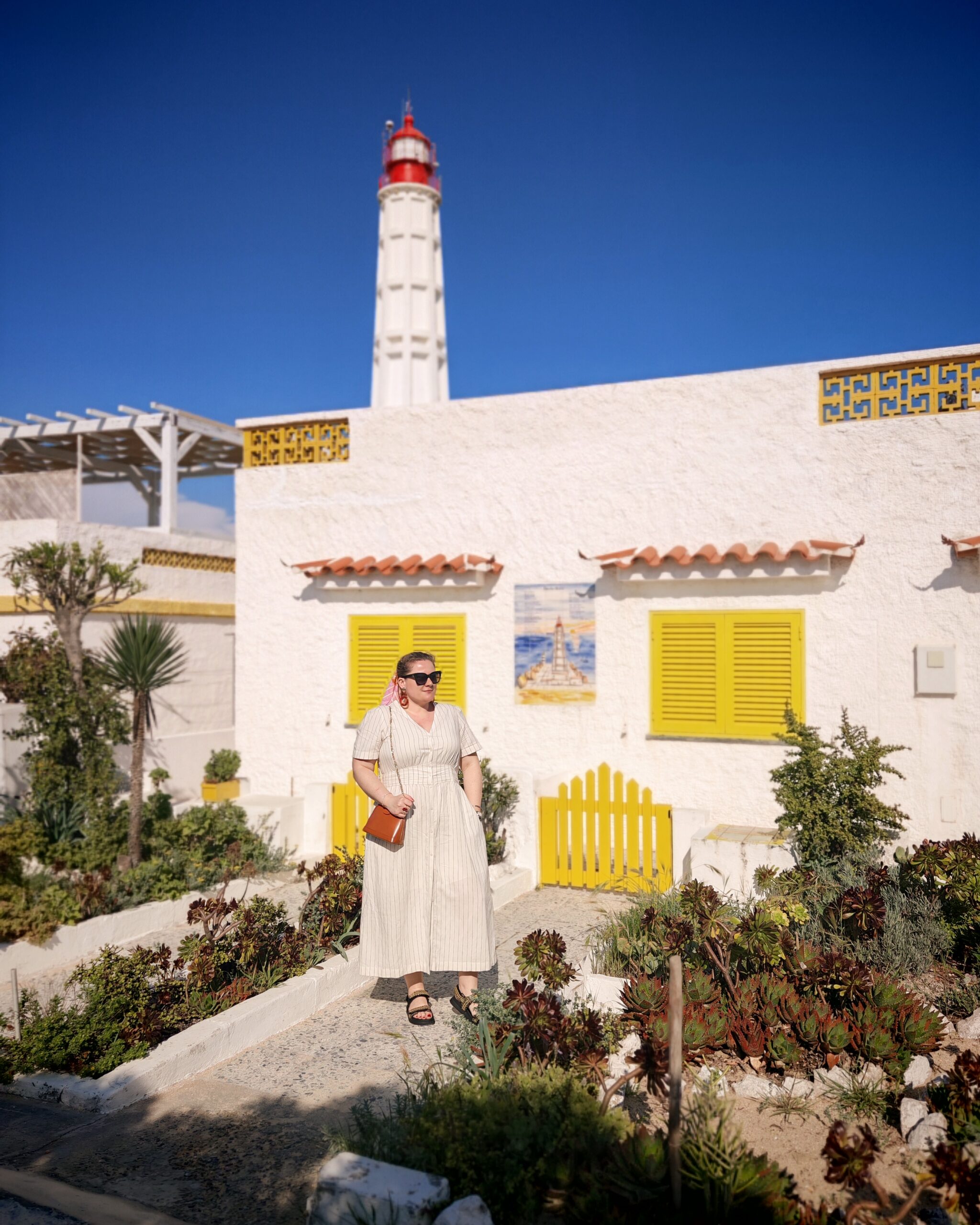 White women wearing a white linen summer dress in front of a white house with yellow shutters and barrier. 