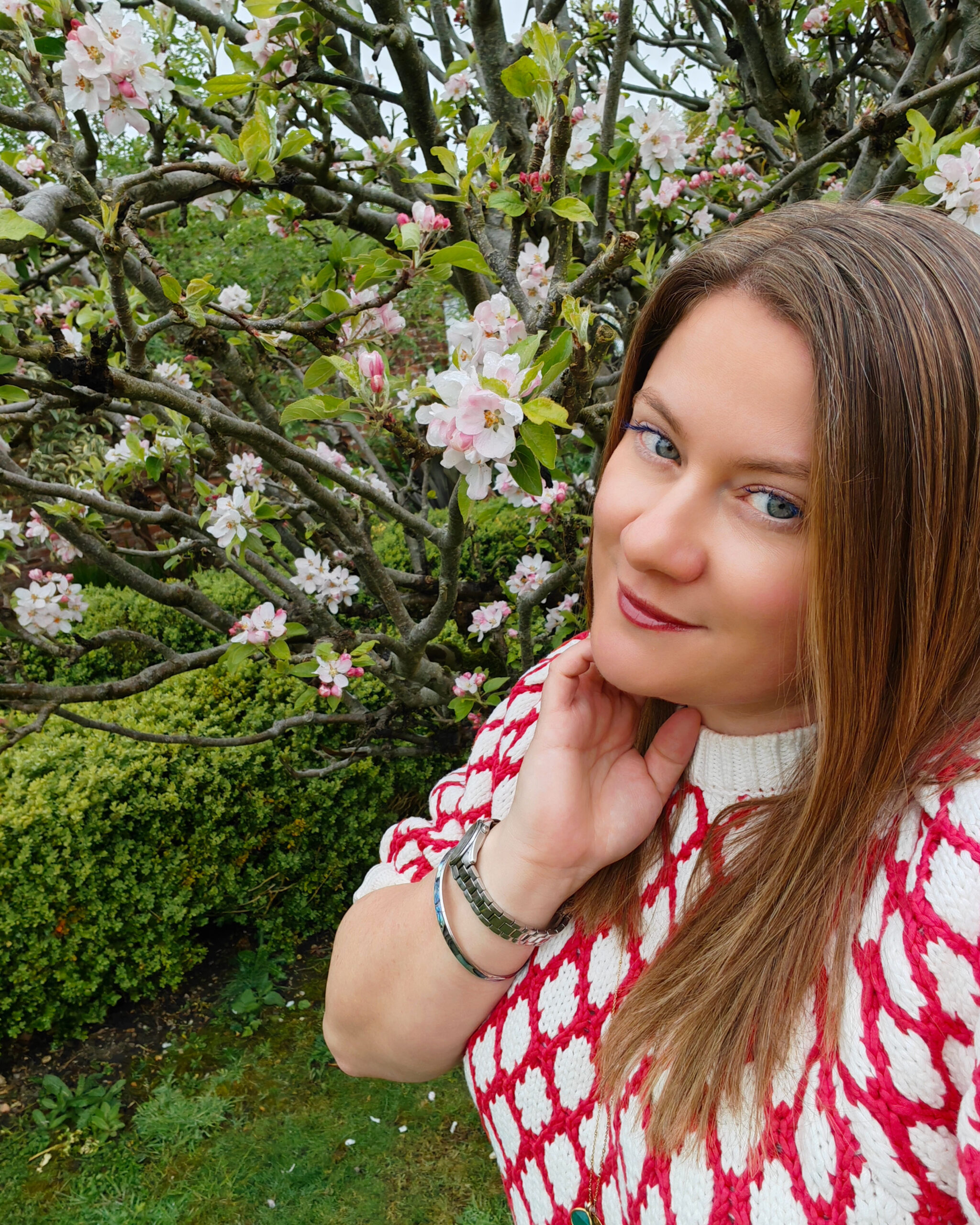 White woman wearing a red and white jumper standing right next to a cherry tree in blossom. She's posing with her hand gently next to her chin.