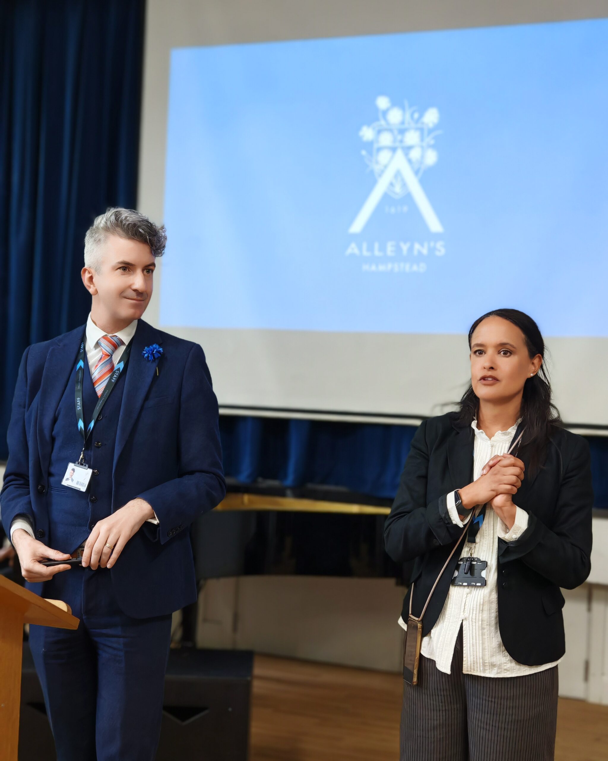 White man wearing a blue suit and mix-raced lady wearing a black suit, giving a presentation during an open day for Alleyn's Hampstead in North London