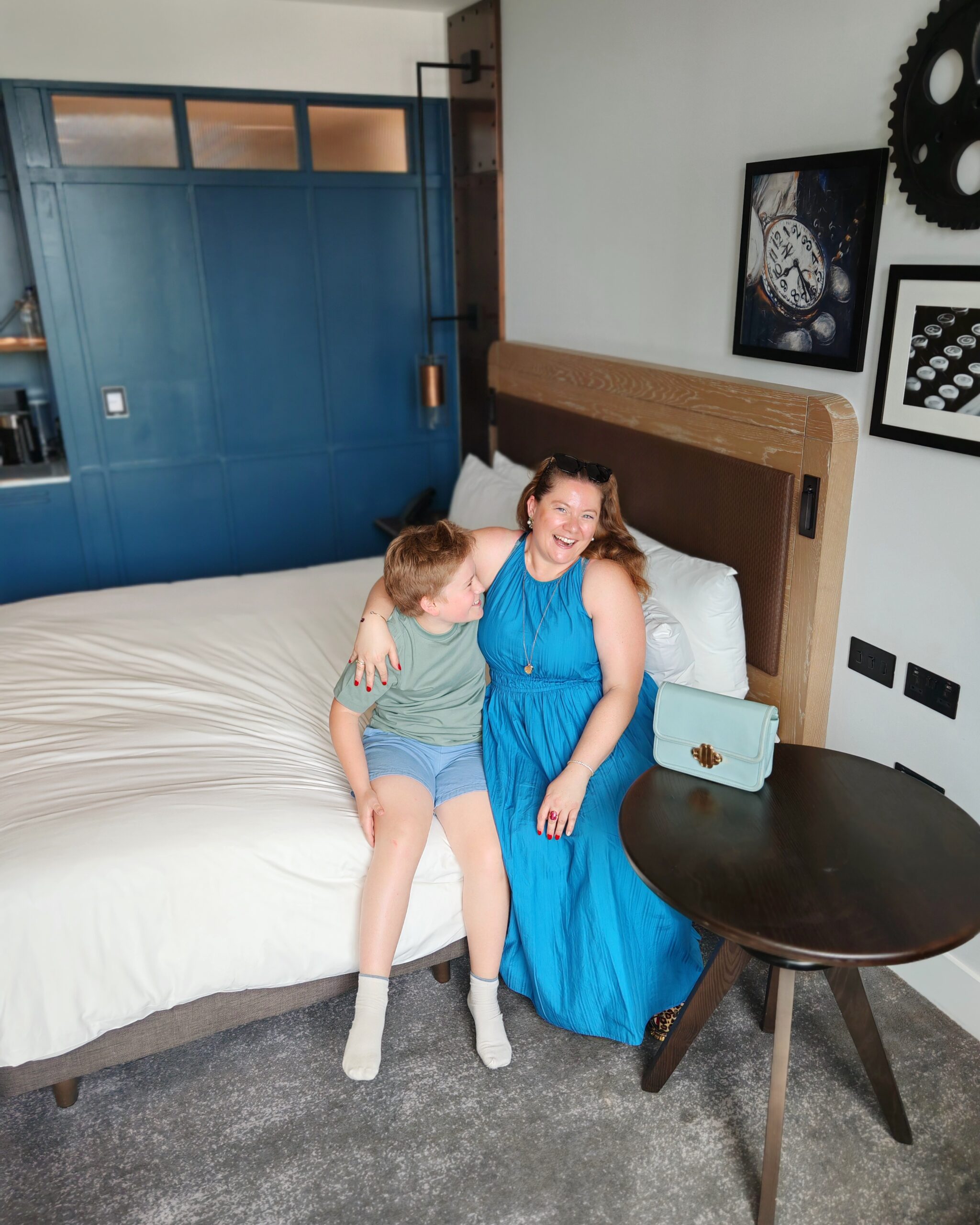 White woman and little boy sitting on a bed in a room from the Gantry Hotel in Stratford, East London. She's laughing at his jokes while wearing a blue dress.