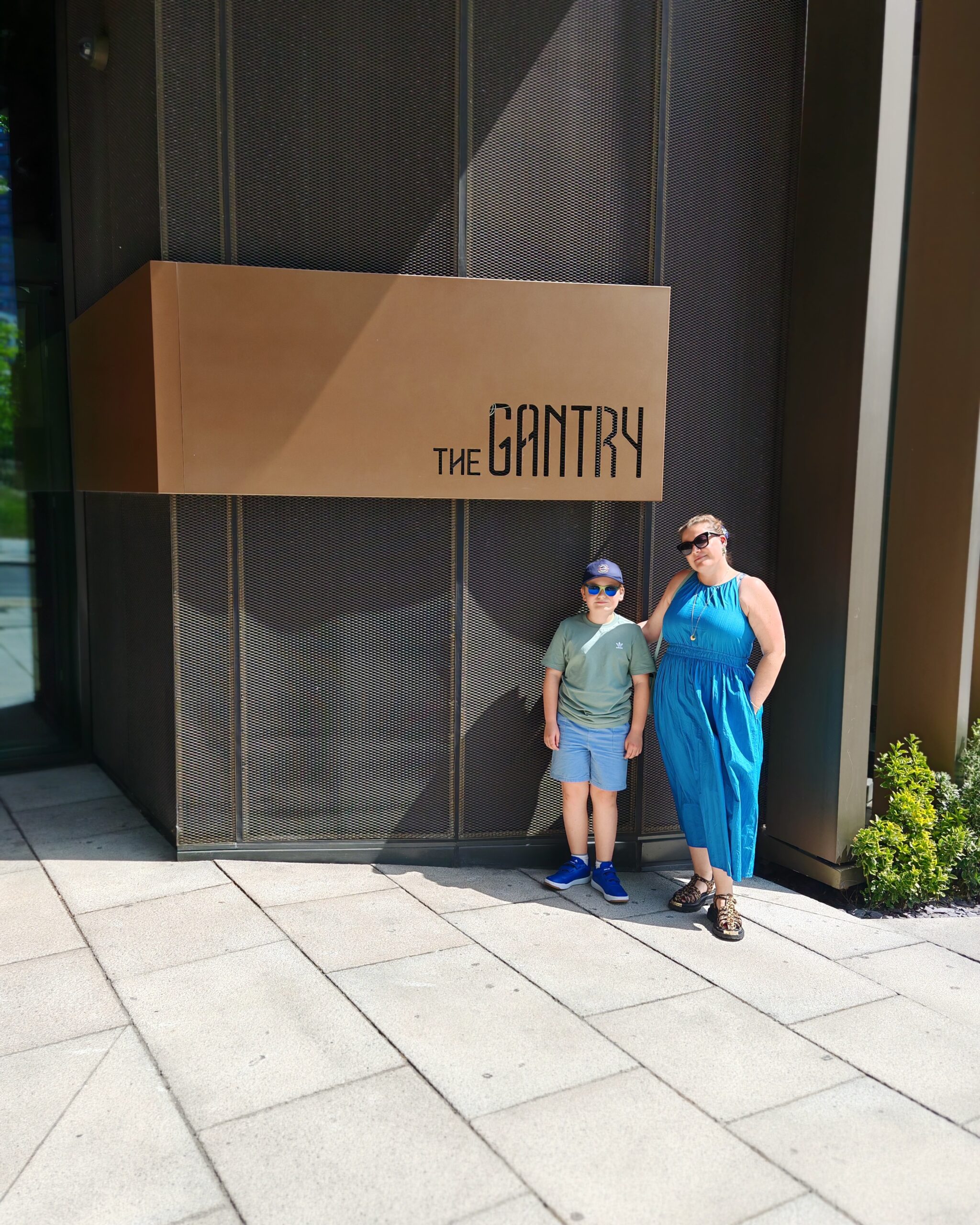 White woman in a blue dress with a white little boy standing up by the entrance of the Gantry Hotel in London.