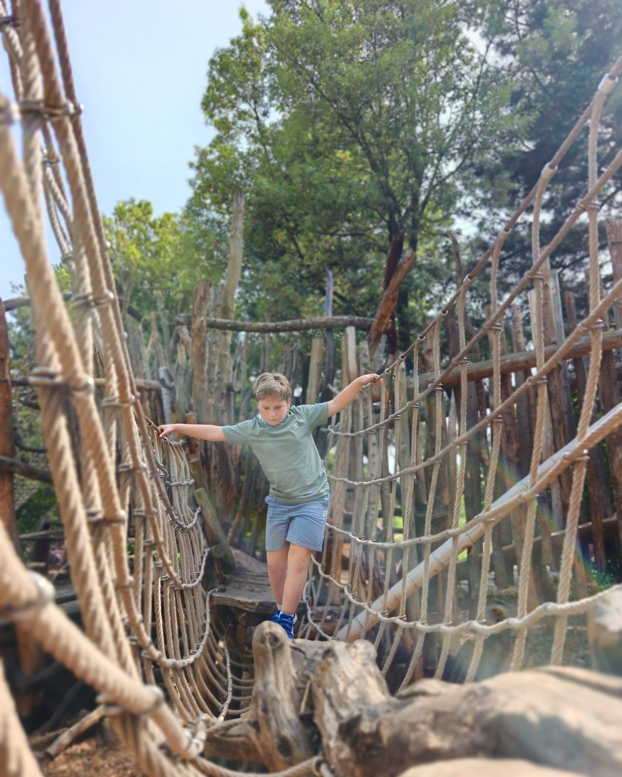 White little boy wearing a green tee and blue shorts playing at the tumbling bay playground in East London, walking along the structure made of ropes