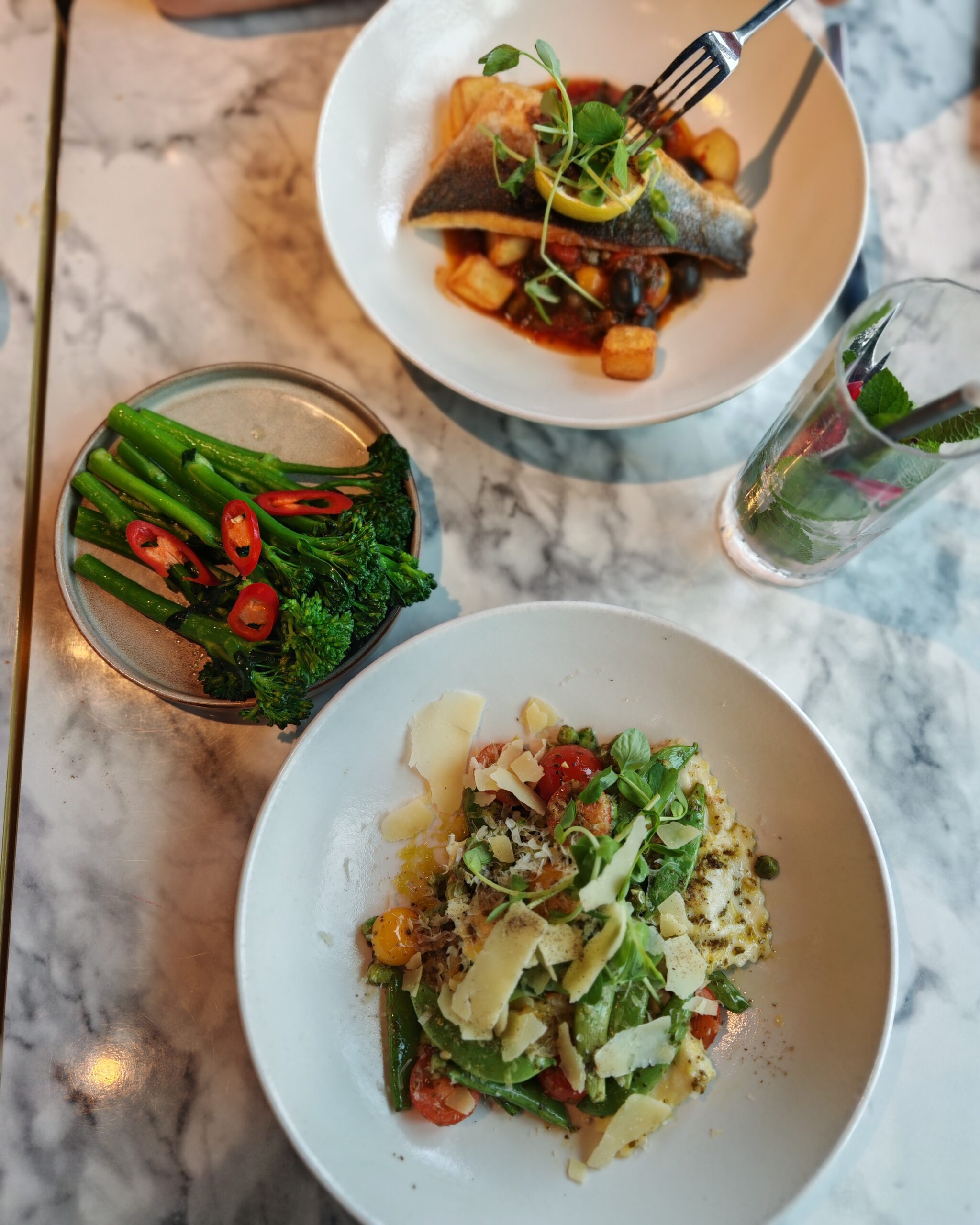 flat lay of food with a side of steam broccoli, seabass at the top and asparagus ravioli at the bottom on a marble like table