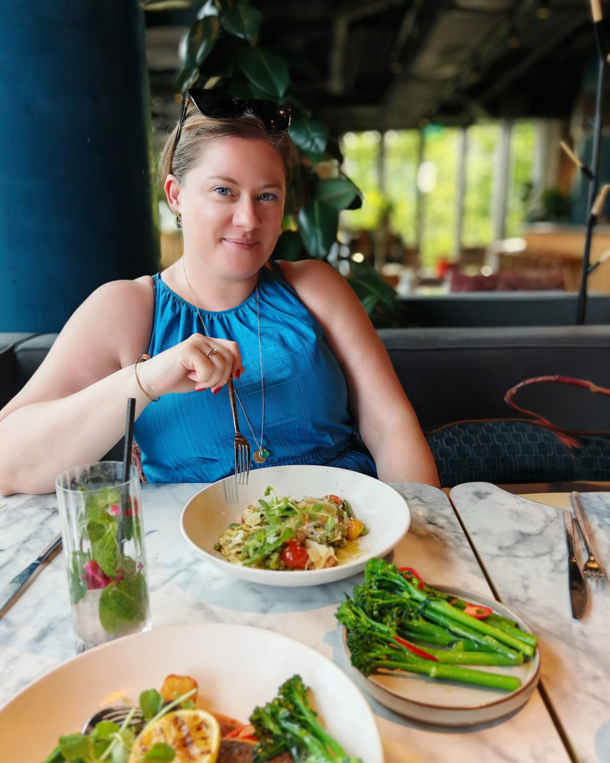 White woman wearing a blue top with a plate of pea ravioli in front of her and some stem broccoli near her plate on a marble table