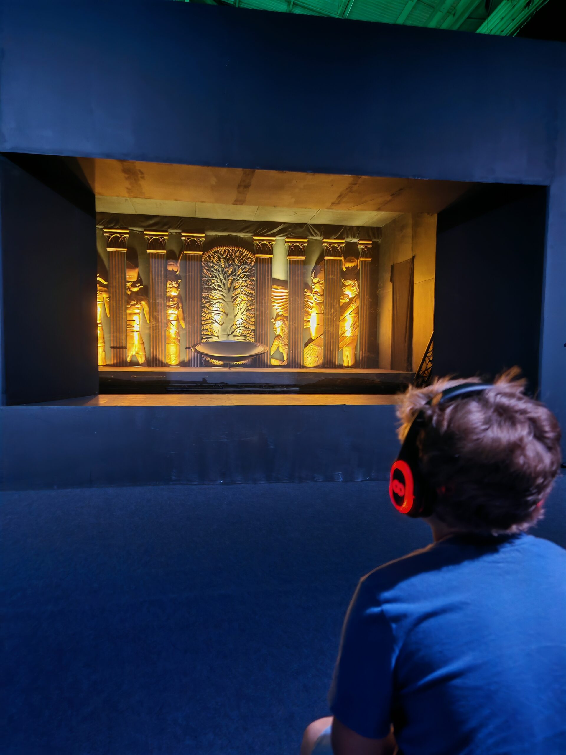 Overlooking the shoulders of a little boy who is looking at a VR exhibition with headphones in London at the Cleopatra Experience at Immerse London
