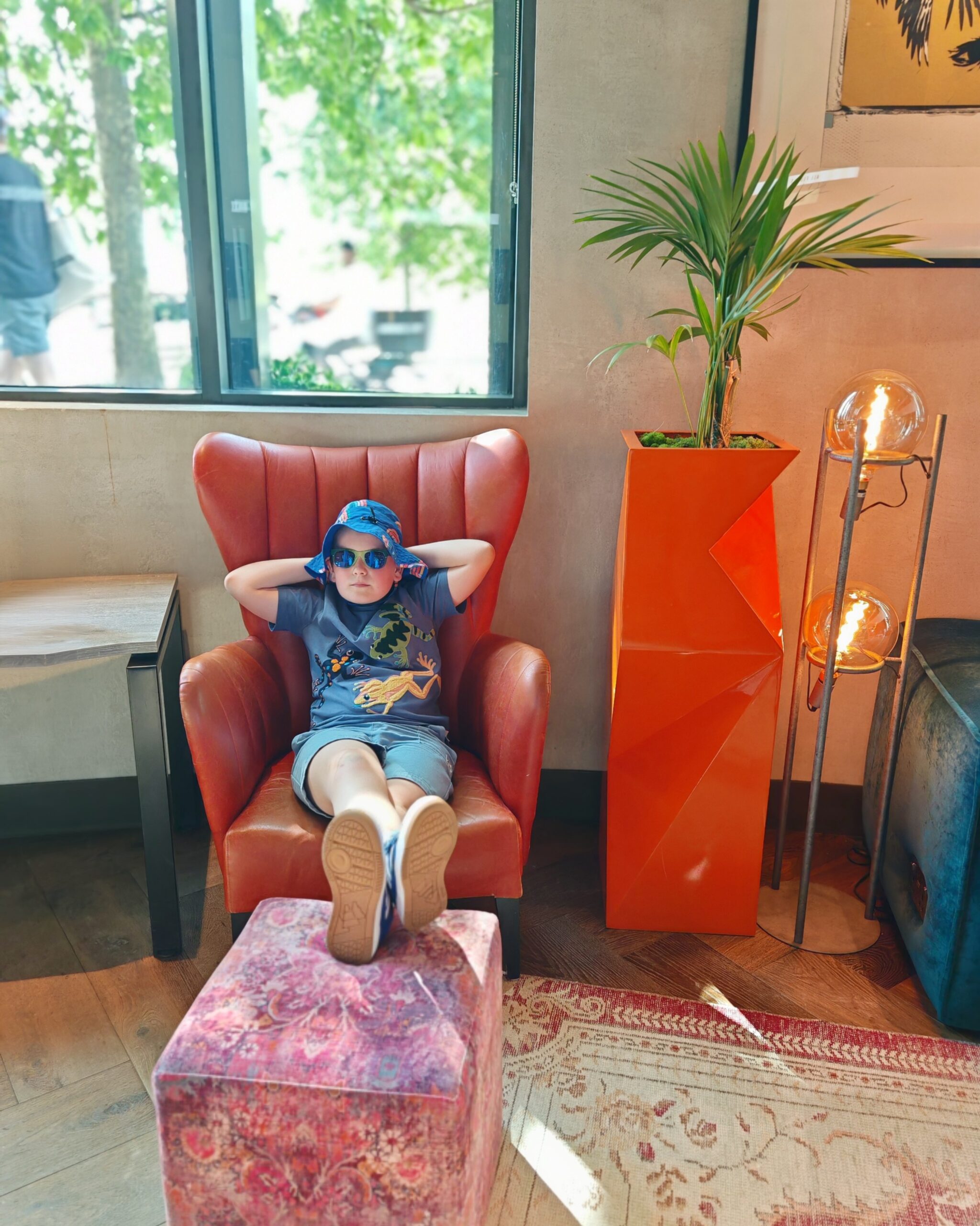 white little boy wearing a blue hat sitting on a leather orange chair in a hotel lobby in London. It's the Gantry Hotel in Stratford.