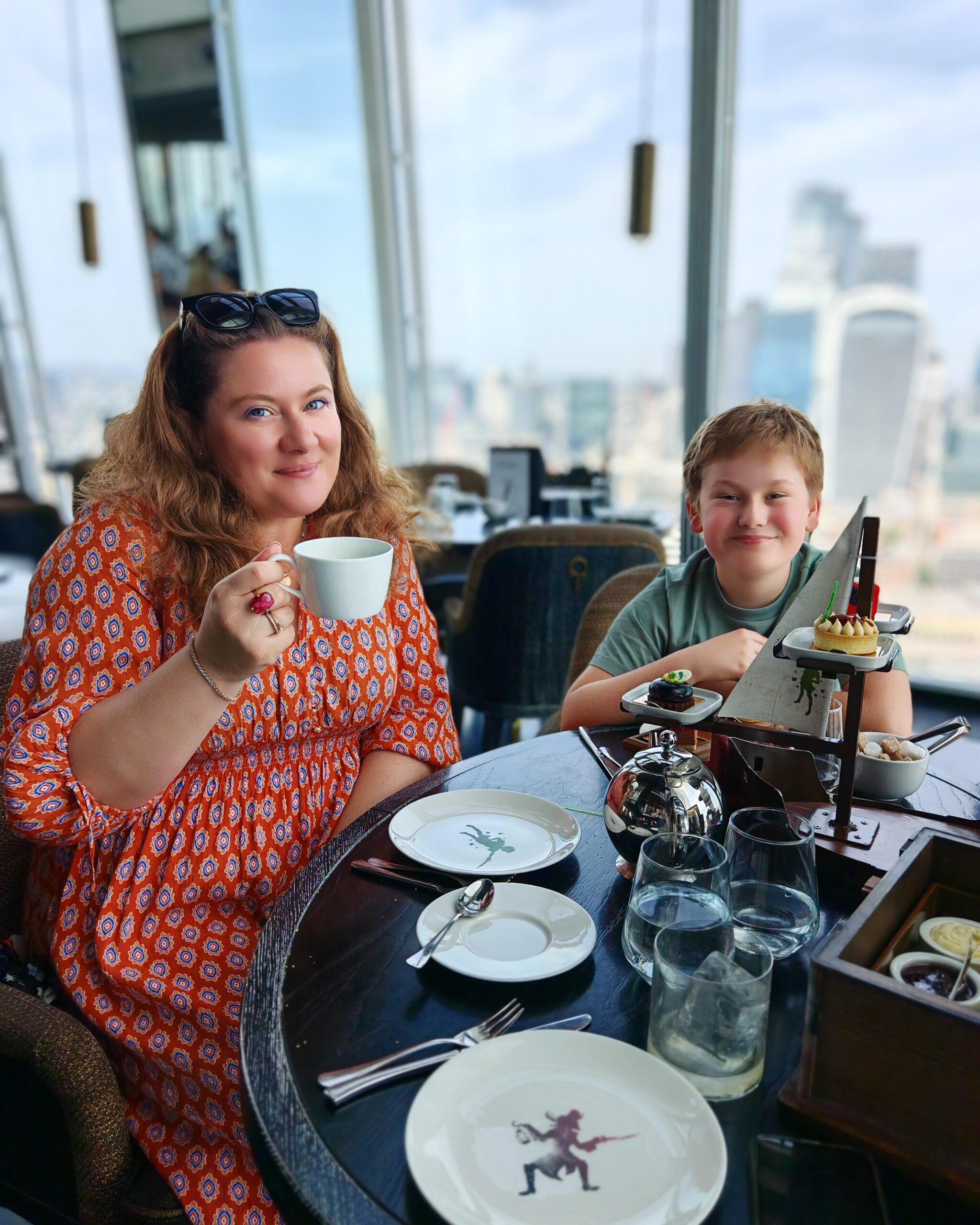 White woman in an orange dress with her son, both smiling and eating the Peter Pan Afternoon Tea at the Aqua Shard restaurant in London White woman in an orange dress with her son, both smiling and eating the Peter Pan Afternoon Tea at the Aqua Shard restaurant in London