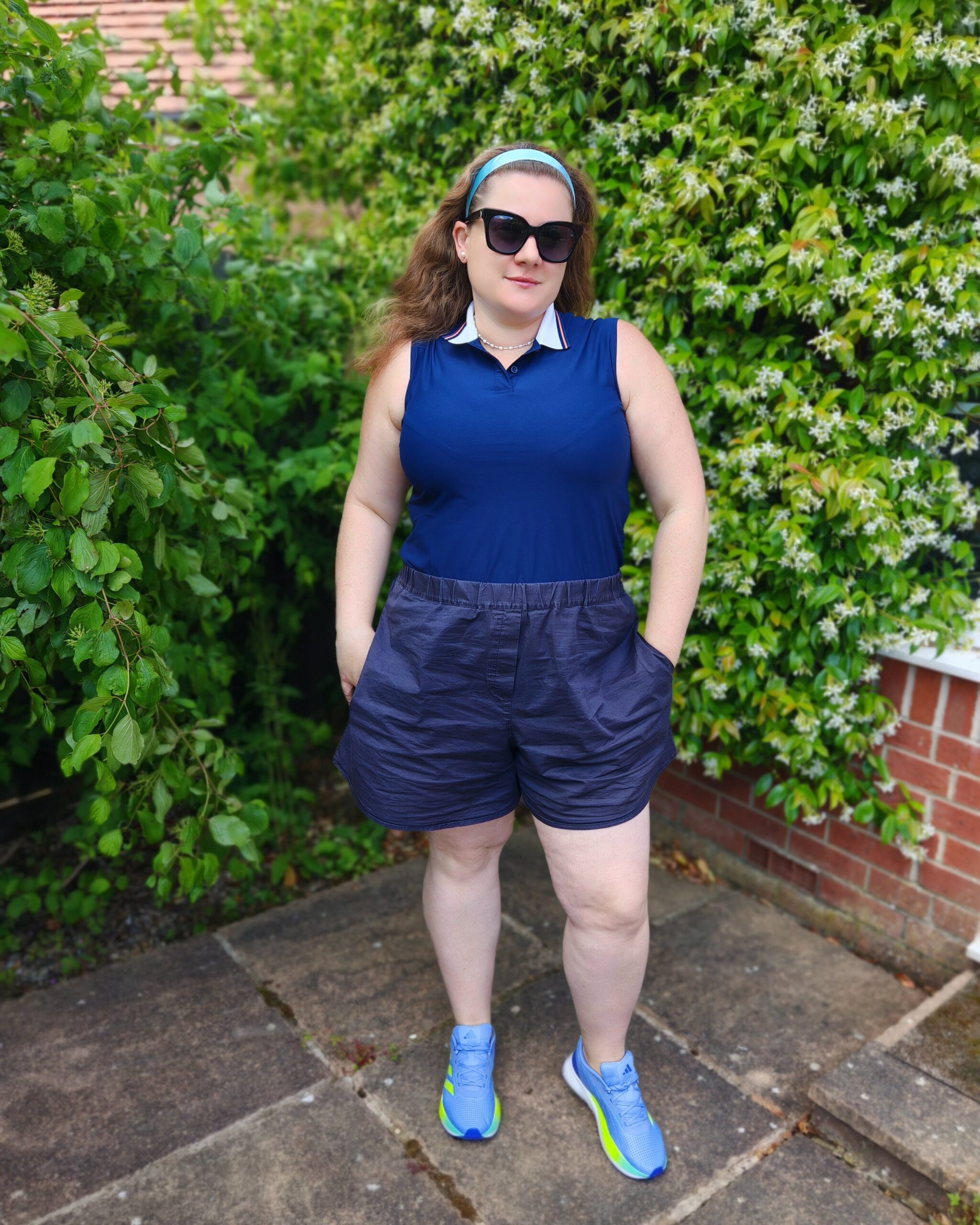 white woman wearing a casual smart outfit for Wimbledon 2026 with a navy adidas polo shirt, some black sunglasses and a navy pair of culottes. She's standing right next to a white flowering shrub white woman wearing a casual smart outfit for Wimbledon 2026 with a navy adidas polo shirt, some black sunglasses and a navy pair of culottes. She's standing right next to a white flowering shrub