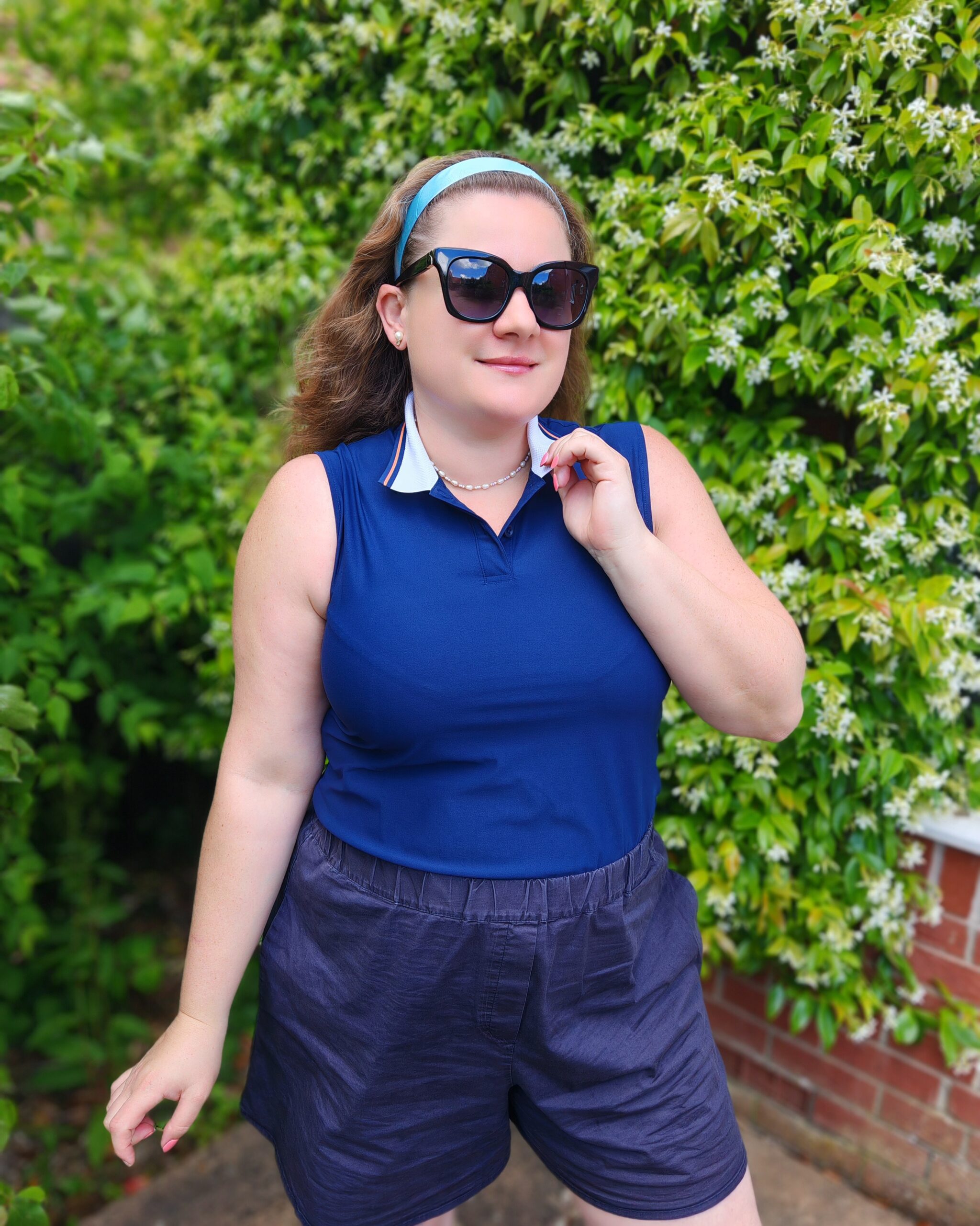white woman wearing a casual smart outfit for Wimbledon 2026 with a navy adidas polo shirt, some black sunglasses and a navy pair of culottes. She's standing right next to a white flowering shrub white woman wearing a casual smart outfit for Wimbledon 2026 with a navy adidas polo shirt, some black sunglasses and a navy pair of culottes. She's standing right next to a white flowering shrub