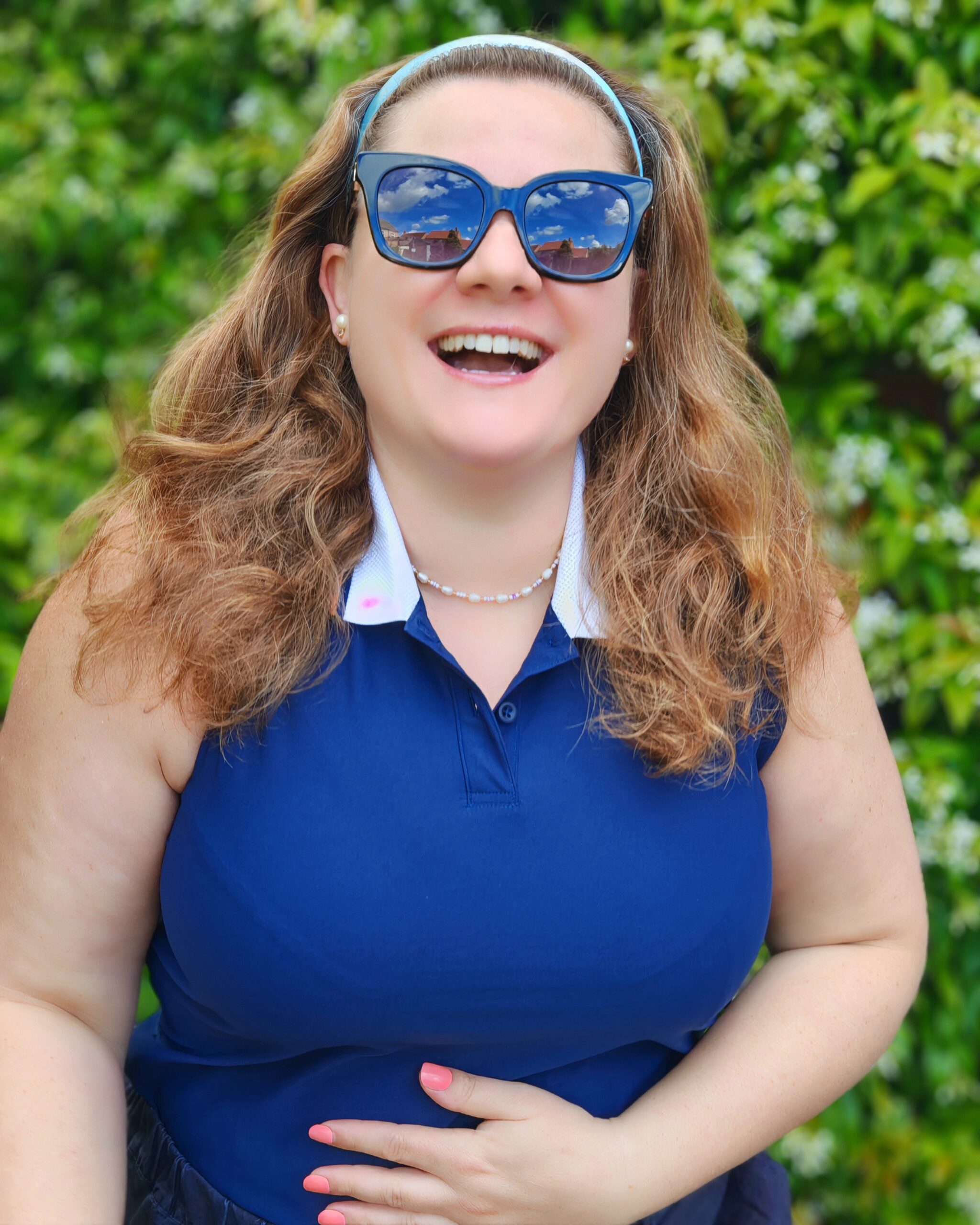 white woman smiling widely while wearing a casual smart outfit for Wimbledon 2026 with a navy adidas polo shirt. She's standing right next to a white flowering shrub white woman smiling widely while wearing a casual smart outfit for Wimbledon 2026 with a navy adidas polo shirt. She's standing right next to a white flowering shrub