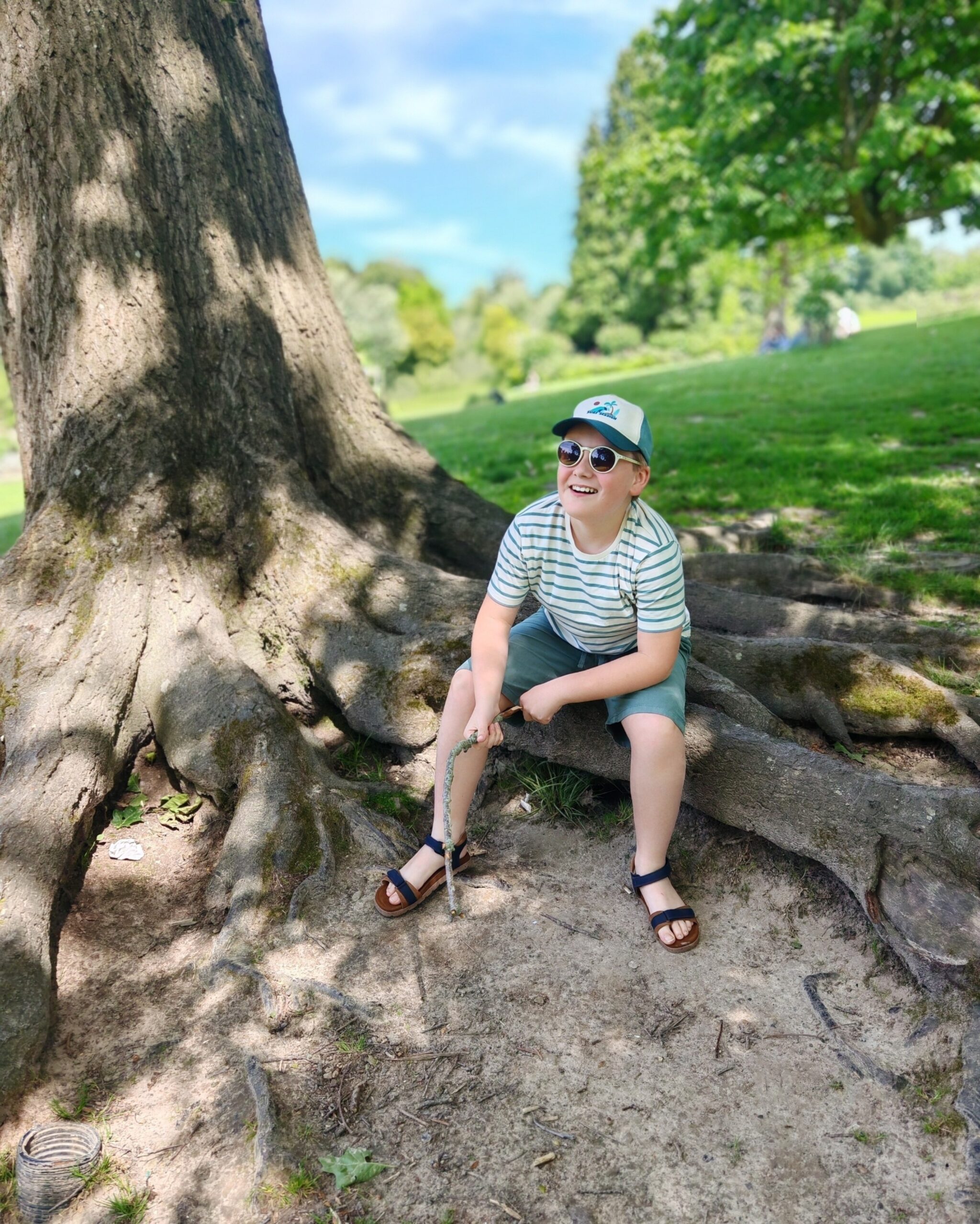A white little boy wearing a cap, a striped tee and sandals. He is sitting on the the roots of a tree in a park on a sunny day