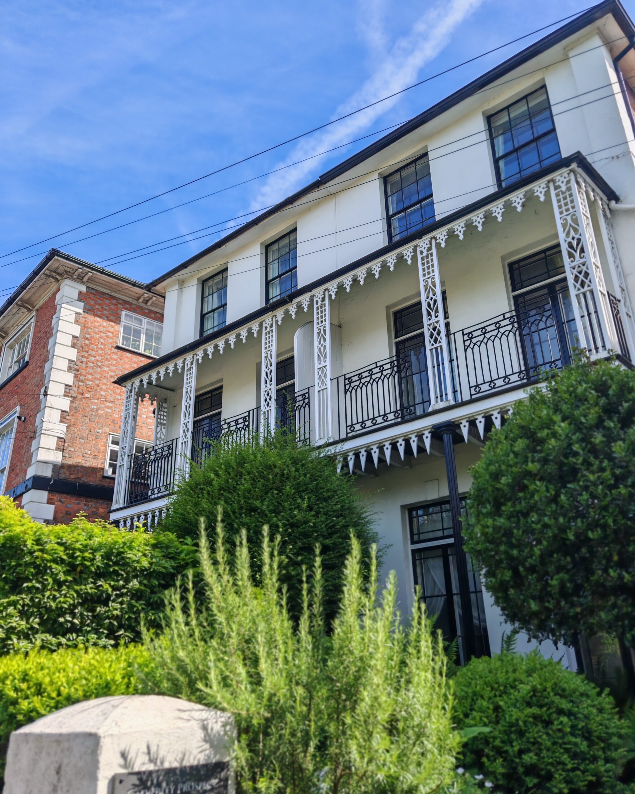 Elegant white Regency-style building with a black wrought iron balcony, intricate white decorative valance, and lush green garden bushes under a bright blue sky