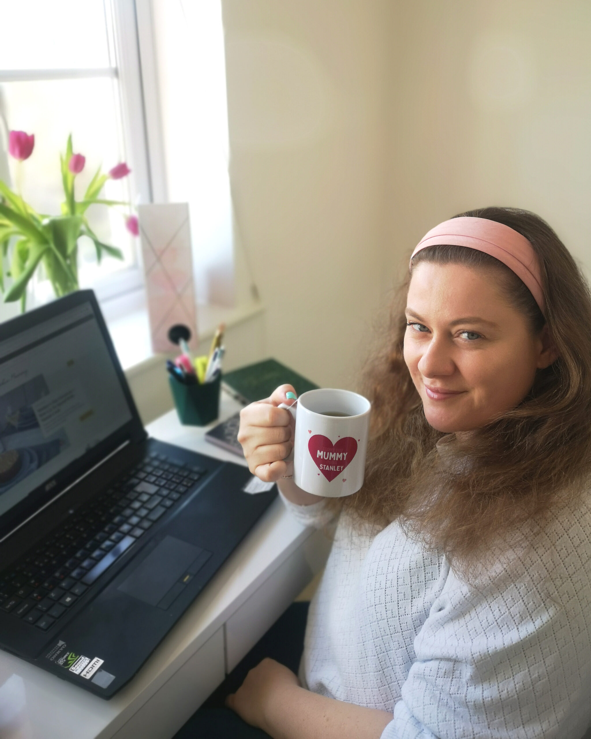White woman in a white jumper, holding a cup of coffee while sitting in front of her laptop to work. She's smiling at the camera White woman in a white jumper, holding a cup of coffee while sitting in front of her laptop to work. She's smiling at the camera
