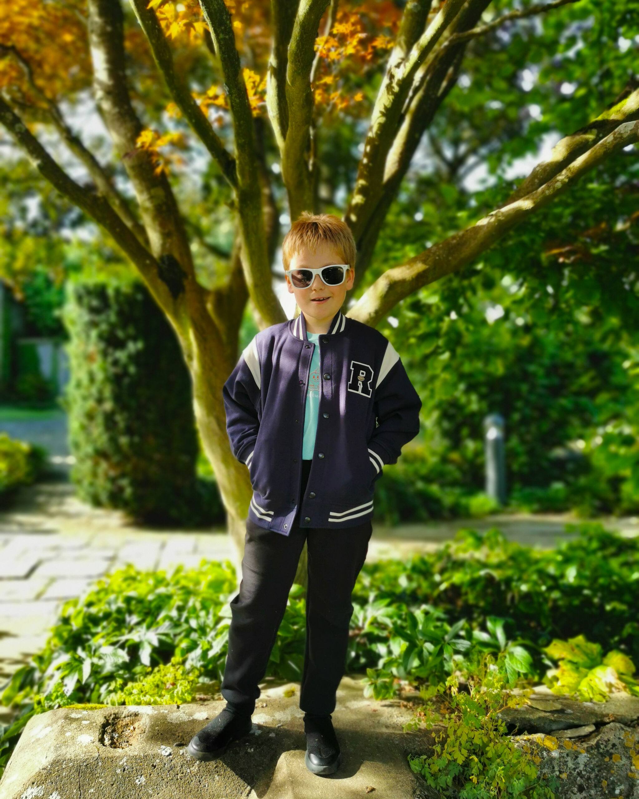 White little boy wearing an American-style bomber jacket posing in a park in Autumn, with brown leaves on the trees
