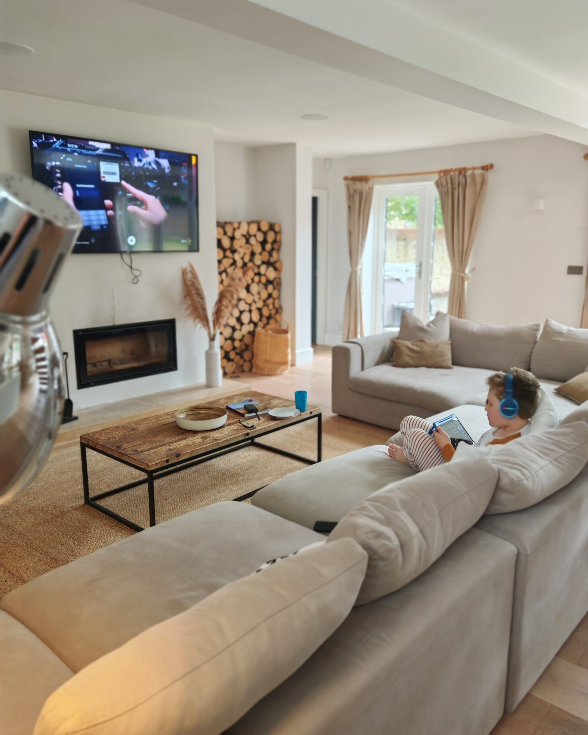 In a large living-room with a large grey sofa and a flat screen with cartoons playing, a little boy is sat while playing on his tablet