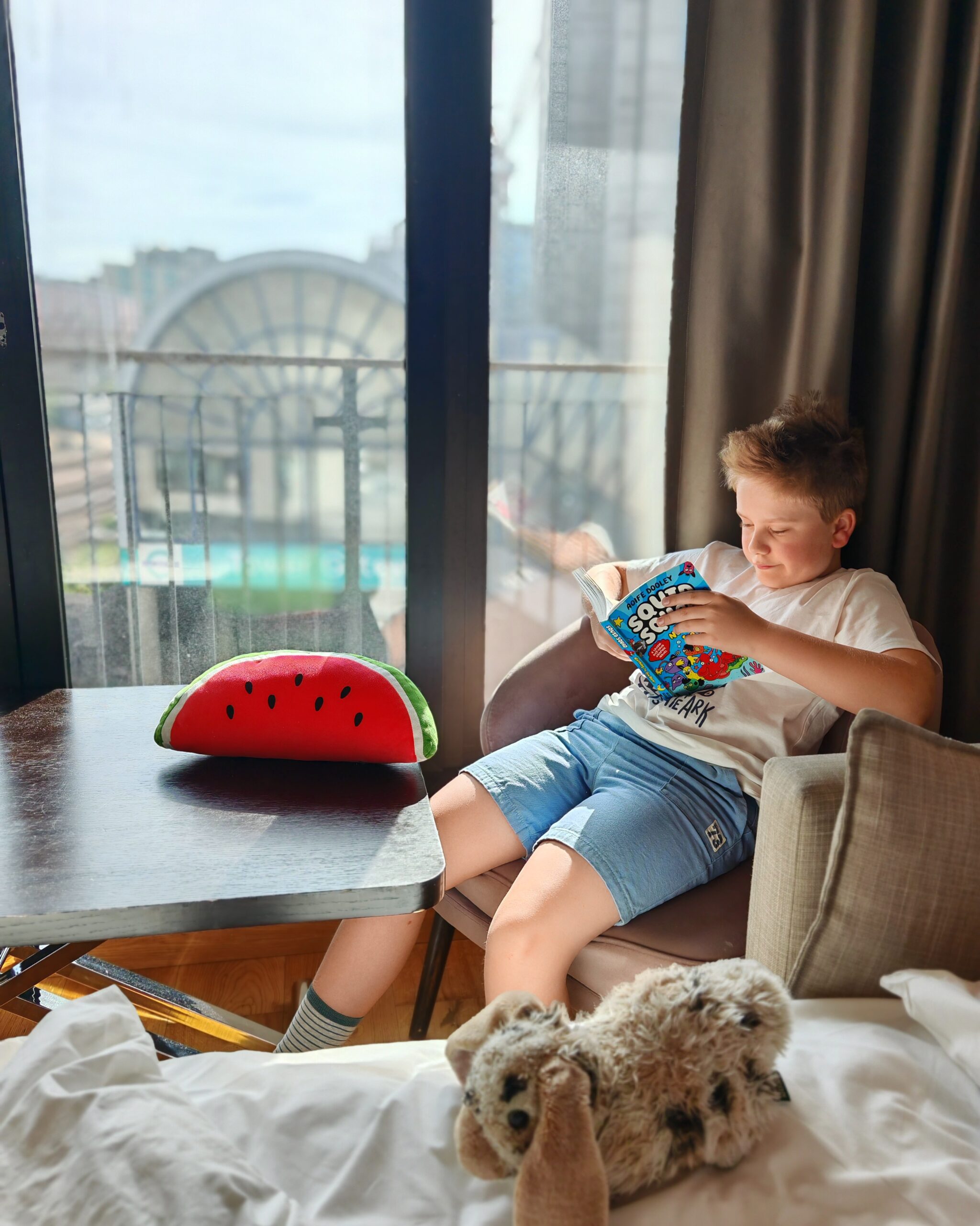 White little boy sitting in a comfy chair in a hotel room in London, right by London Blackfriars station. He's reading a book with a watermelon soft toy on the table next to him White little boy sitting in a comfy chair in a hotel room in London, right by London Blackfriars station. He's reading a book with a watermelon soft toy on the table next to him