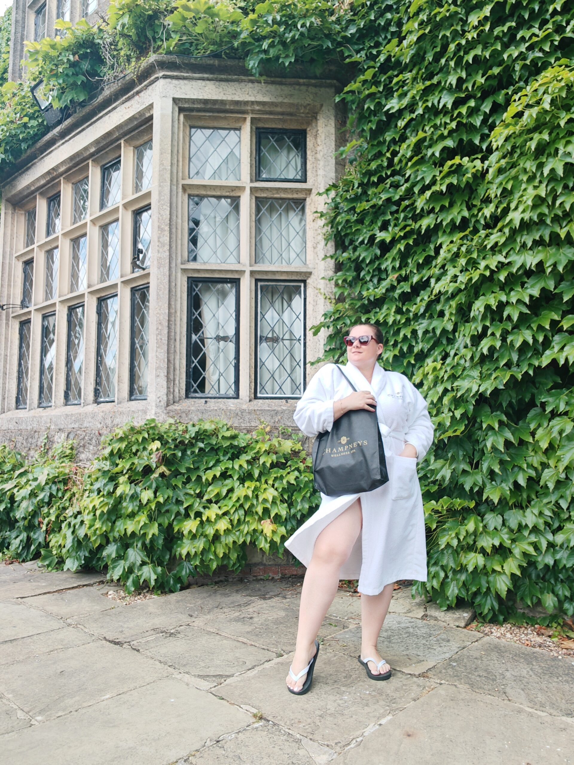 White woman in a robe standing right next to a window at Eastwell Manor Champneys Spa in Kent. She's holding a black back from the spa hotel while looking ahead of her
