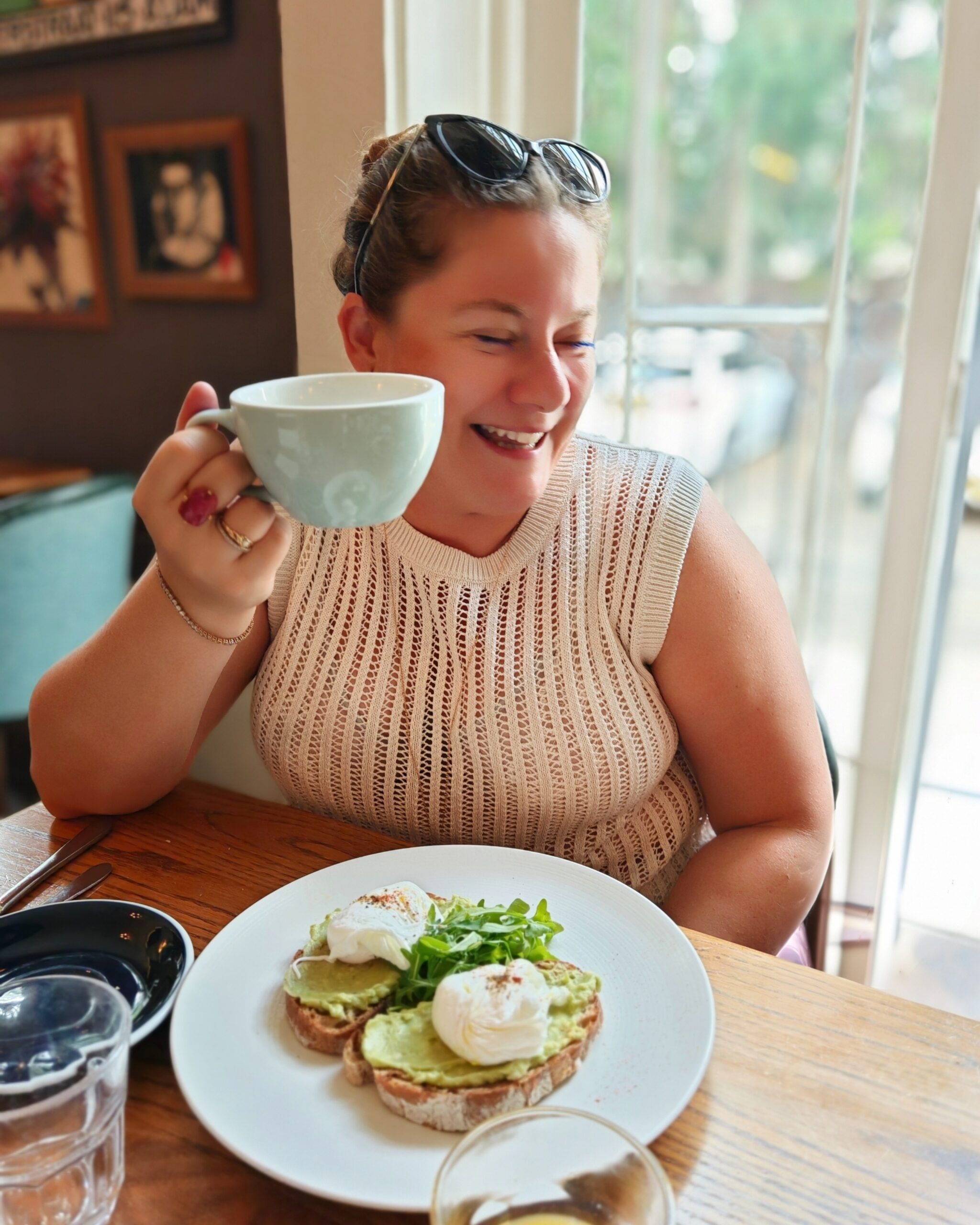 White woman wearing a beige crochet top smiling while eating smashed avocado on toast with poached eggs.