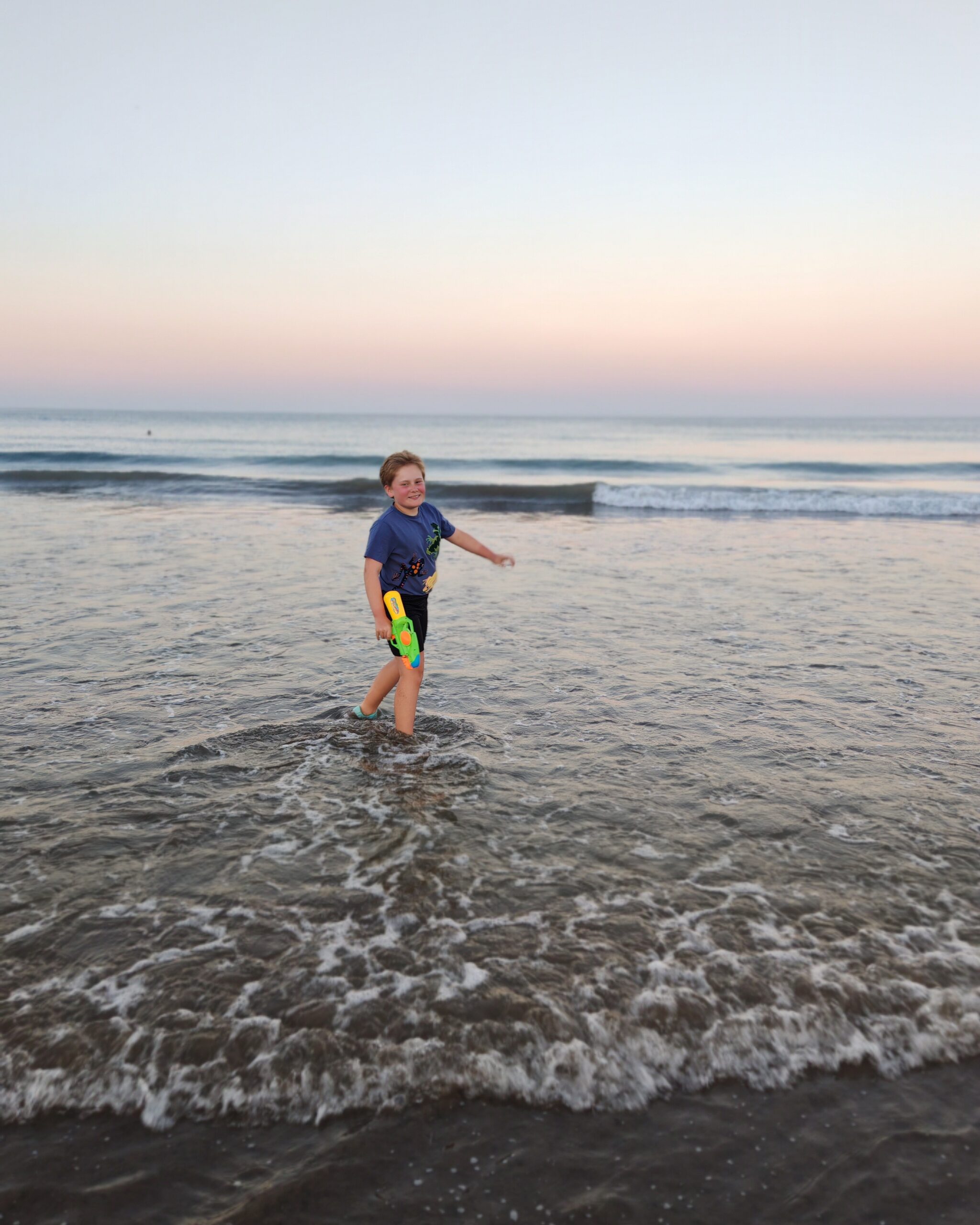 White little boy in a blue tee-shirt and black shorts with a water gun, playing in the water at la Playa del inglés in Gran Canaria