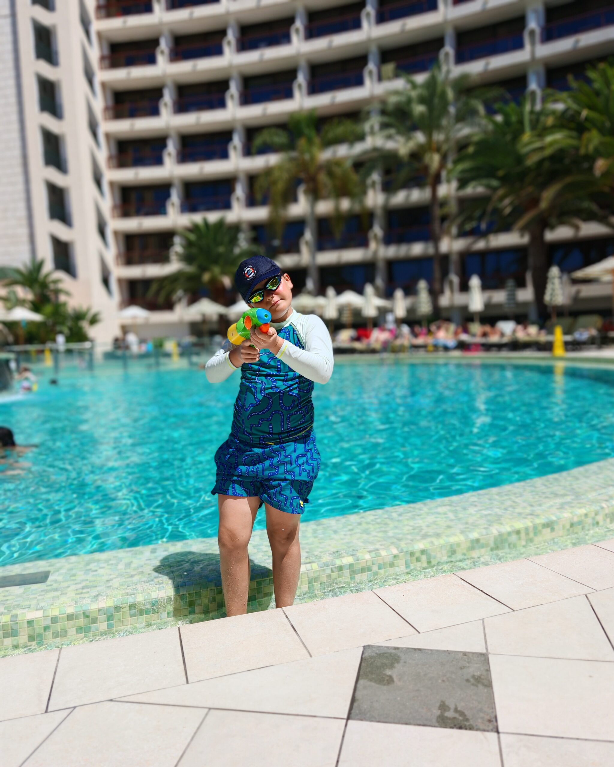 white little boy in a blue cap and swimsuit with a water gun by the pool at the Seaside Sandy Beach Hotel