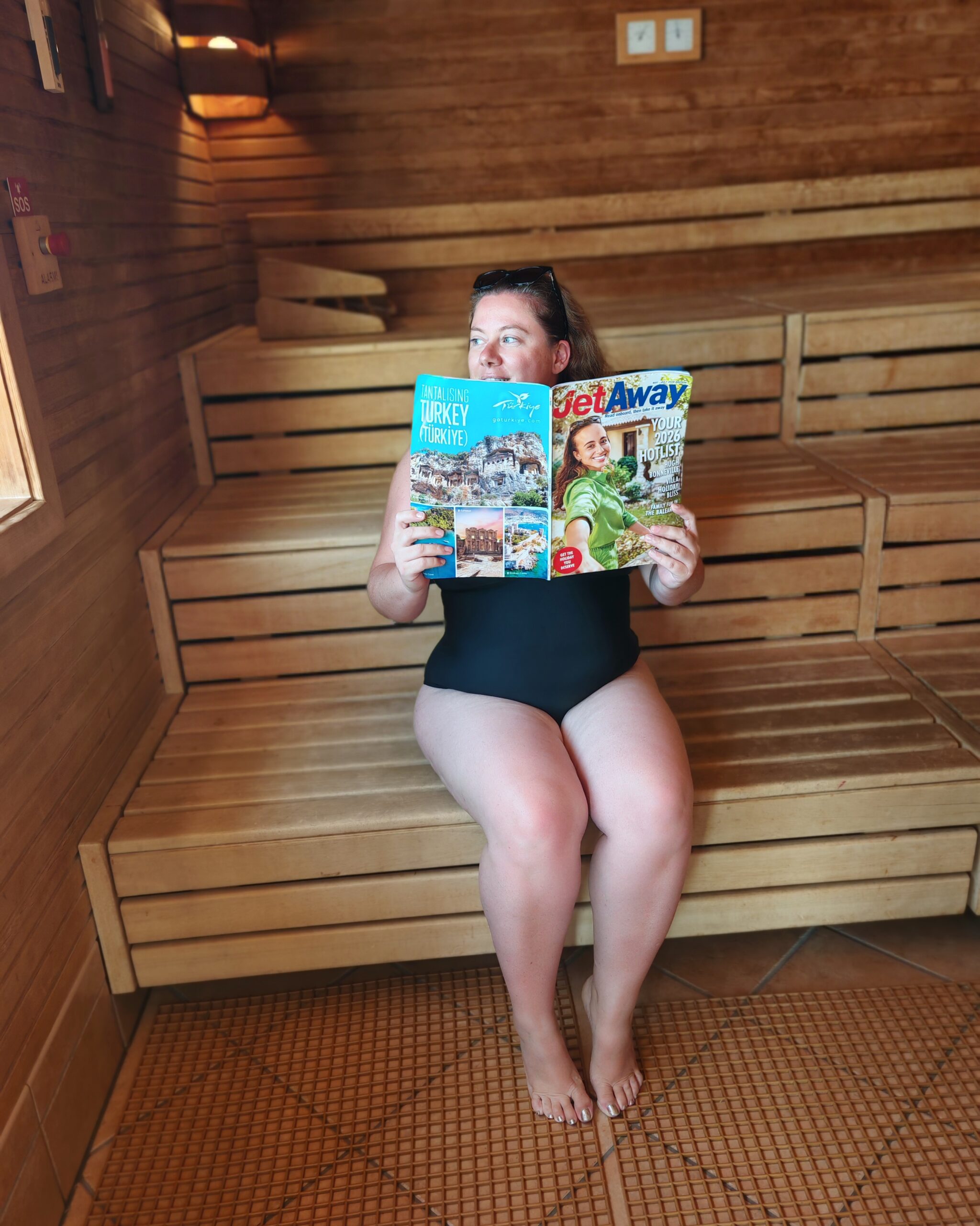 white woman in a black swimming costume, reading the JetAway Magazine while sitting in the sauna at the spa