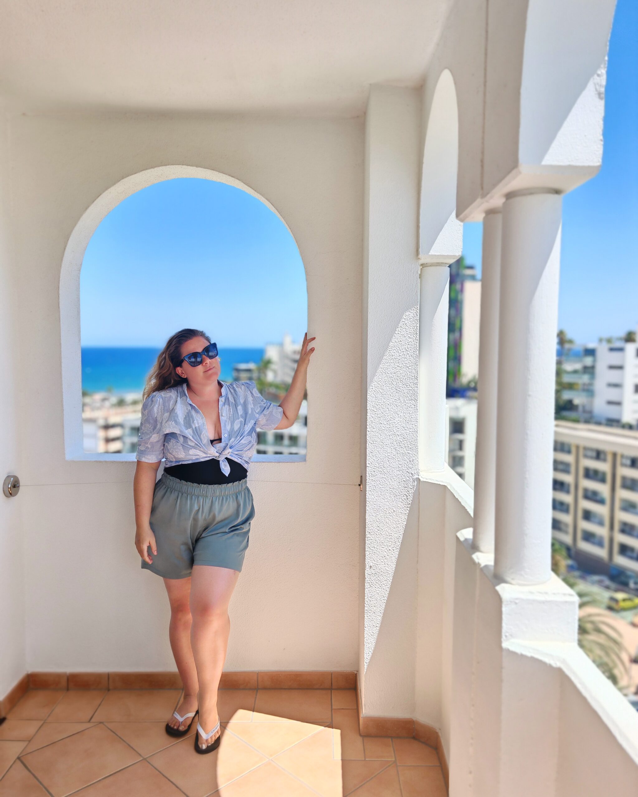 White woman in a blue top and shorts, standing on the balcony at the Seaside Sandy Beach Hotel