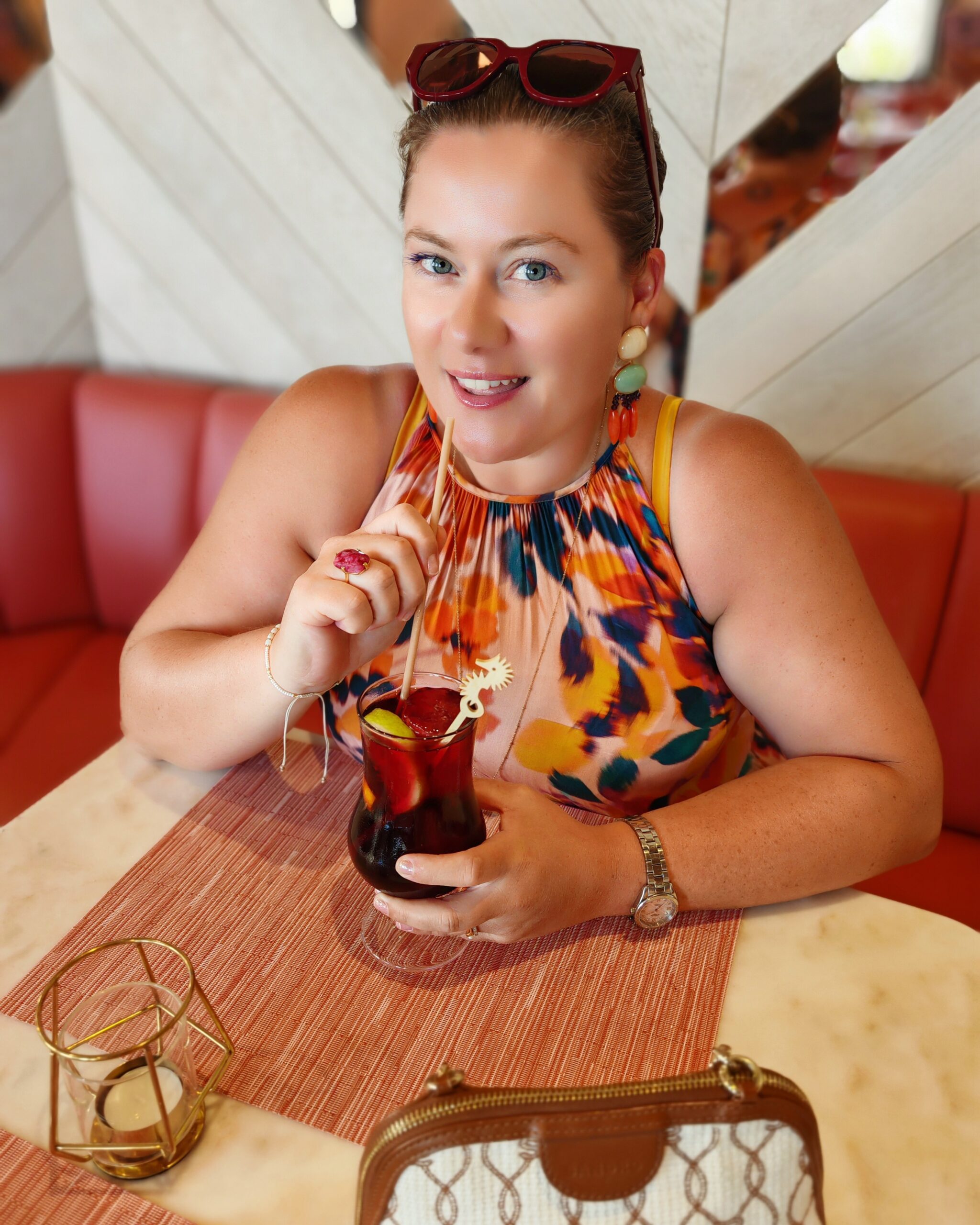 White woman in a pink dress with glowing skin at a pink bar on holiday. She's smiling to the camera, holding a sangria she's about to drink