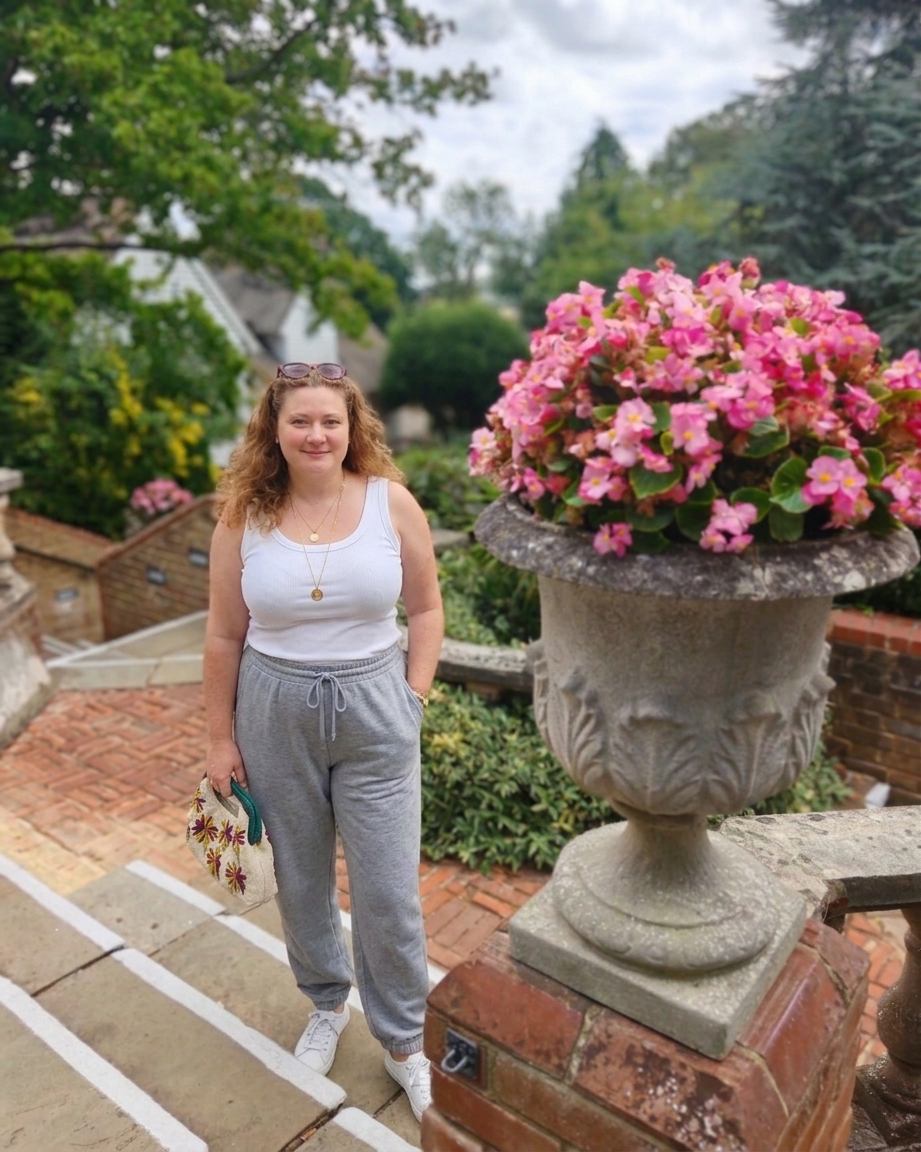 White woman in a white top tank and a pair of grey sweatpants standing by a big pot of pink flowers at Rowhill Grange in Kent