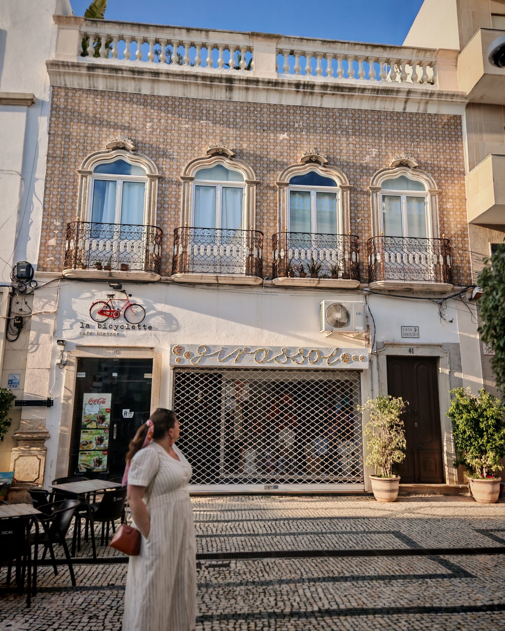 White woman in a beige linen dress standing in front of an European shop. It could be Italy or Spain