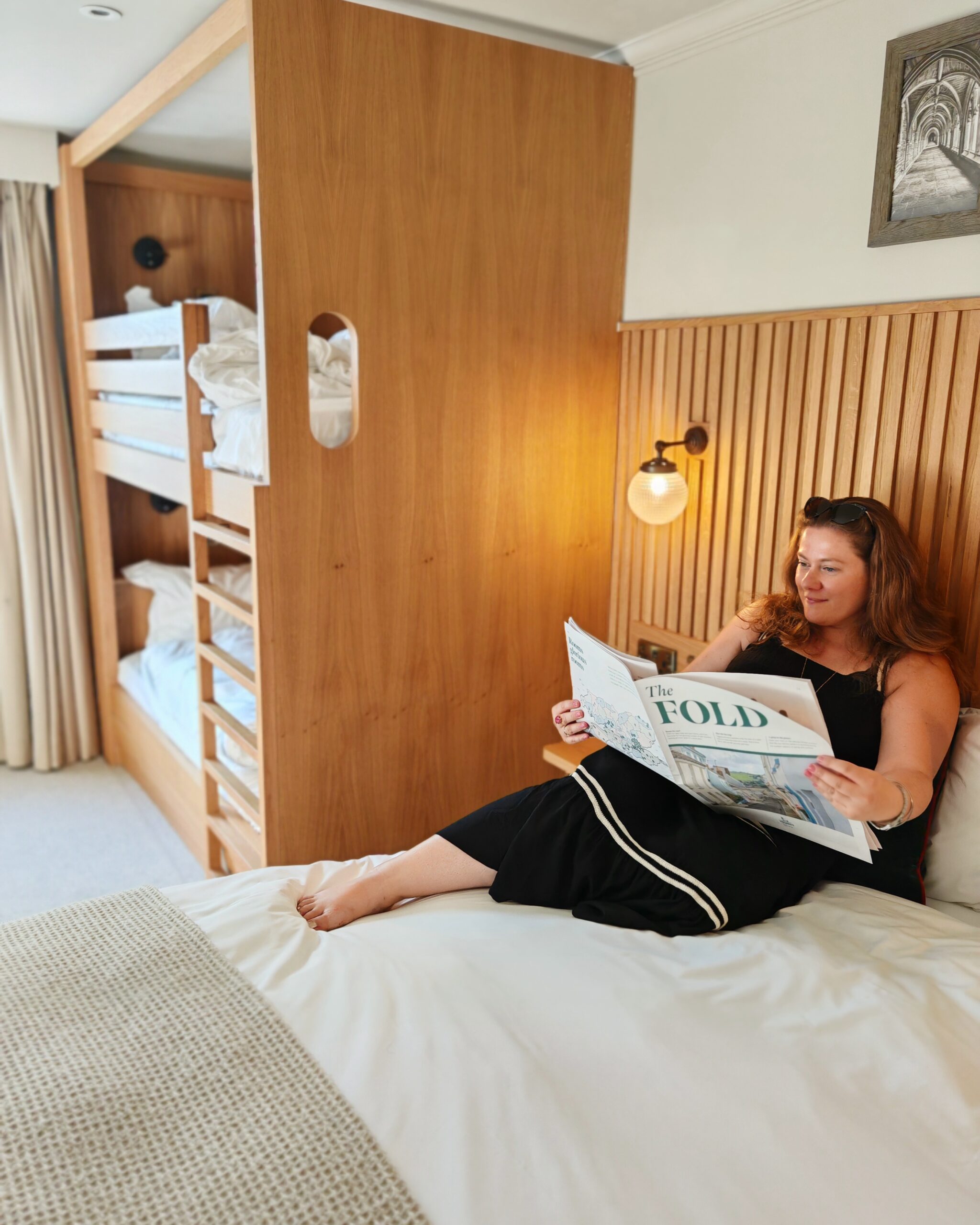 White woman in a black mid-dress seating on a bed and reading the Fold magazine at the Park Teddington in South-West London