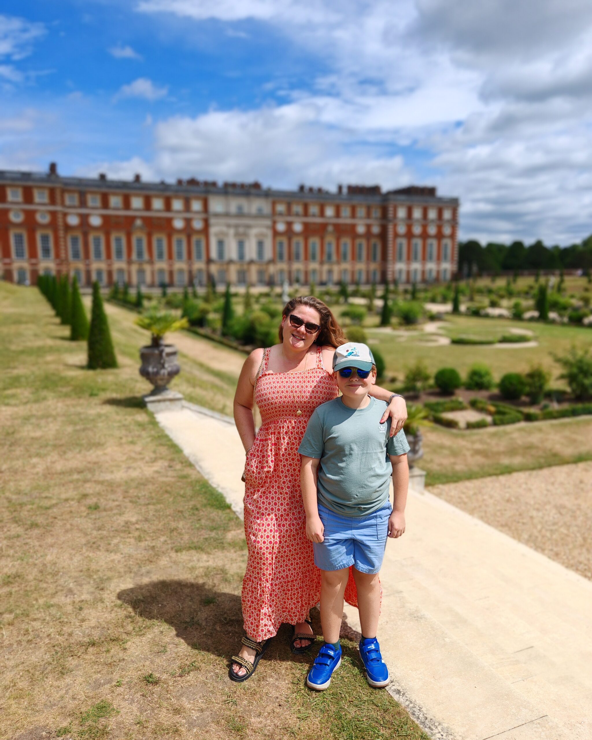 White woman in an orange long dress with her son posing in front of Hampton Court Palace side gardens
