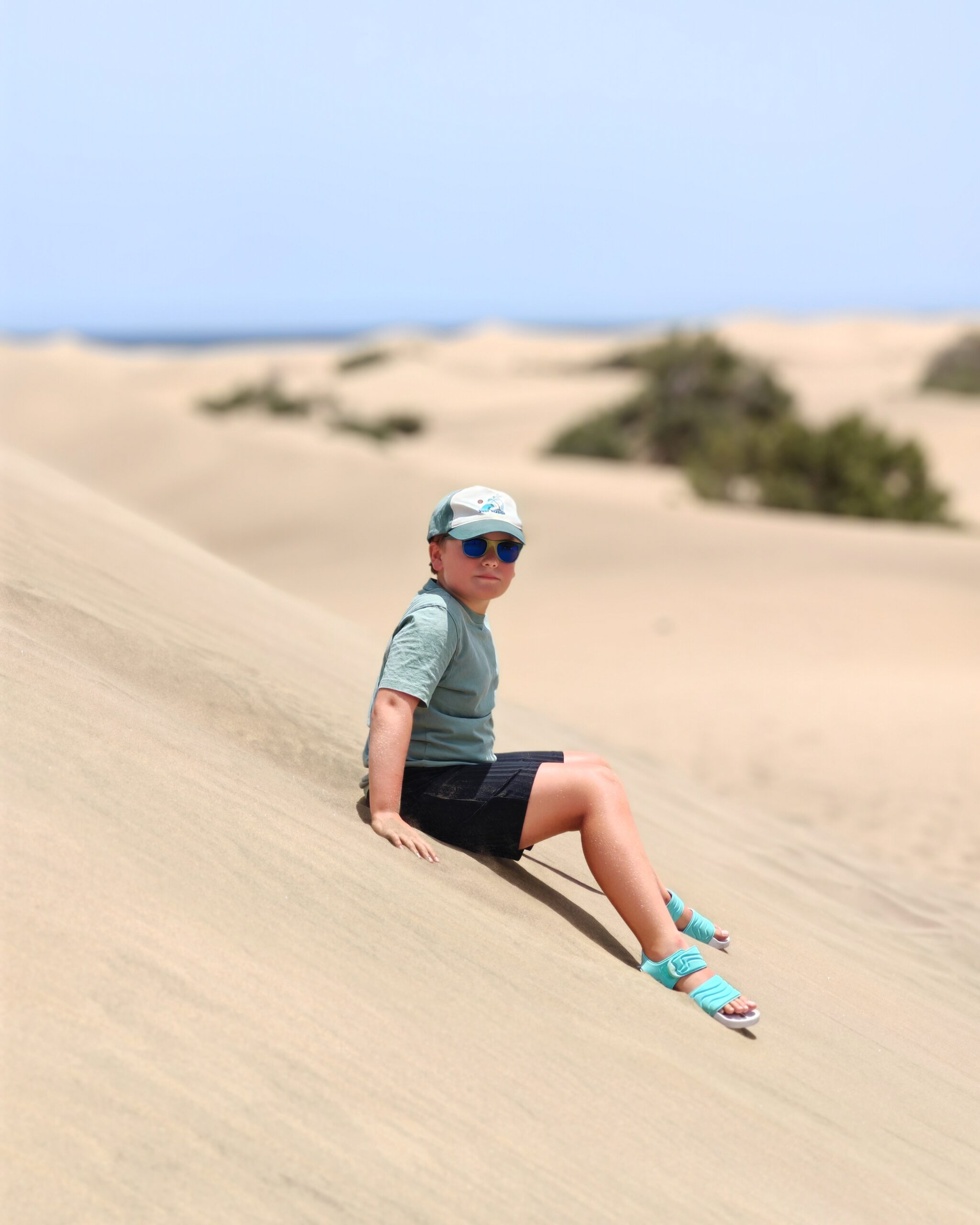 White little boy wearing black shorts, a green tee and a white cap in the dunes of Maspalomas in the sunshine