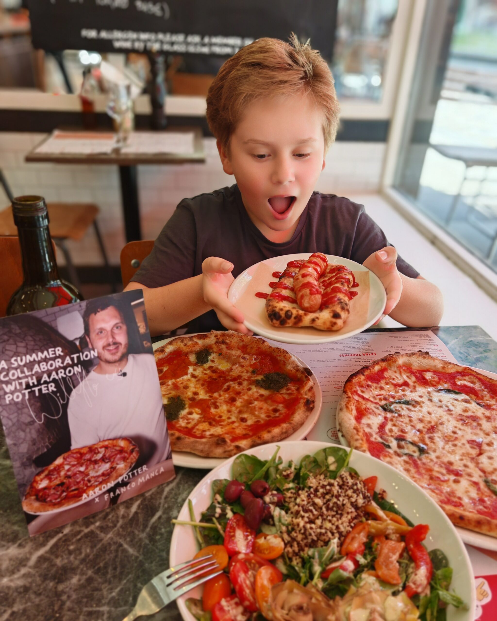 White little boy at Franco Manca, holding a plate and looking like he's going to eat the new hot dog from the kids' summer menu