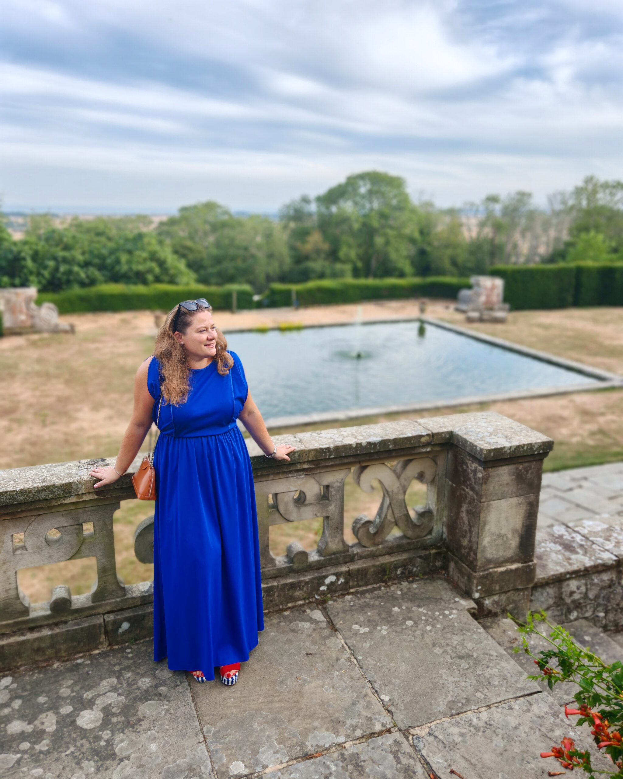 White woman in a long vintage blue dress standing on the balcony at Port Lympne House right by the fountain White woman in a long vintage blue dress standing on the balcony at Port Lympne House right by the fountain