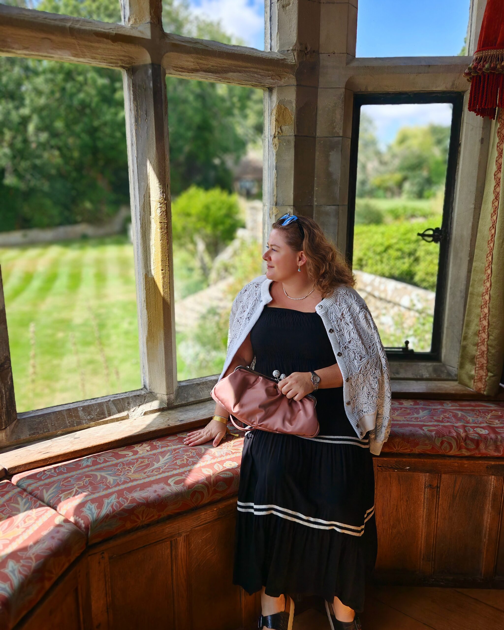 White woman in a black dress and a beige bomber jacket sat on the window edge of Lympne Castle. She's overlooking at the garden outside White woman in a black dress and a beige bomber jacket sat on the window edge of Lympne Castle. She's overlooking at the garden outside