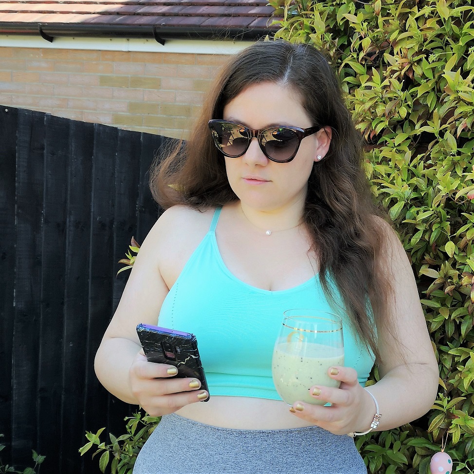 White woman with long chesnut hair and green sports bra looking at he phone while drinking a green smoothie White woman with long chesnut hair and green sports bra looking at he phone while drinking a green smoothie