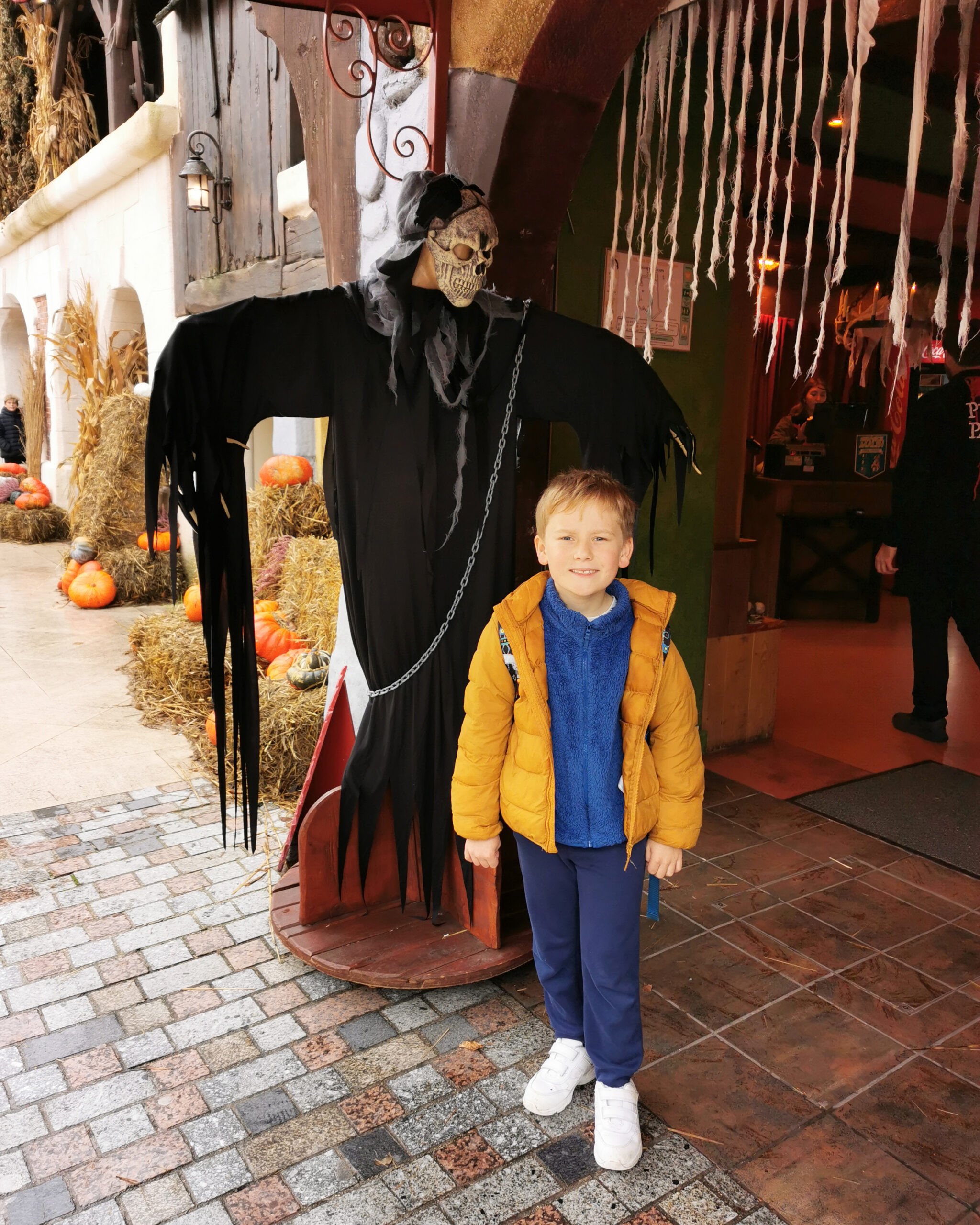 white little boy in a blue outfit and a yellow parka standing in front of a ghost statue at Parc Asterix in Paris, France