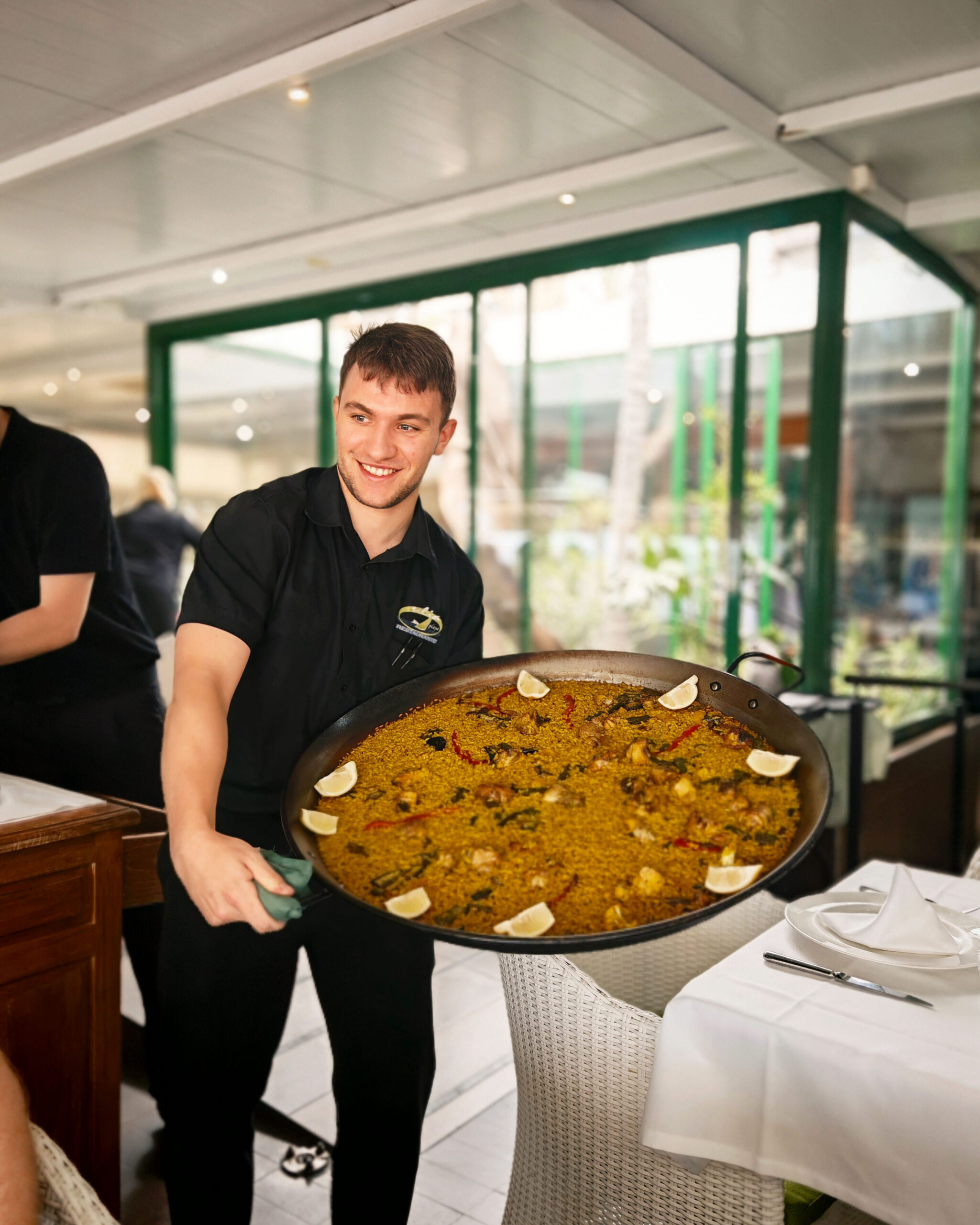 Spanish waiter all in black holding a massive plate of seafood paella in a restaurant in Benidorm.