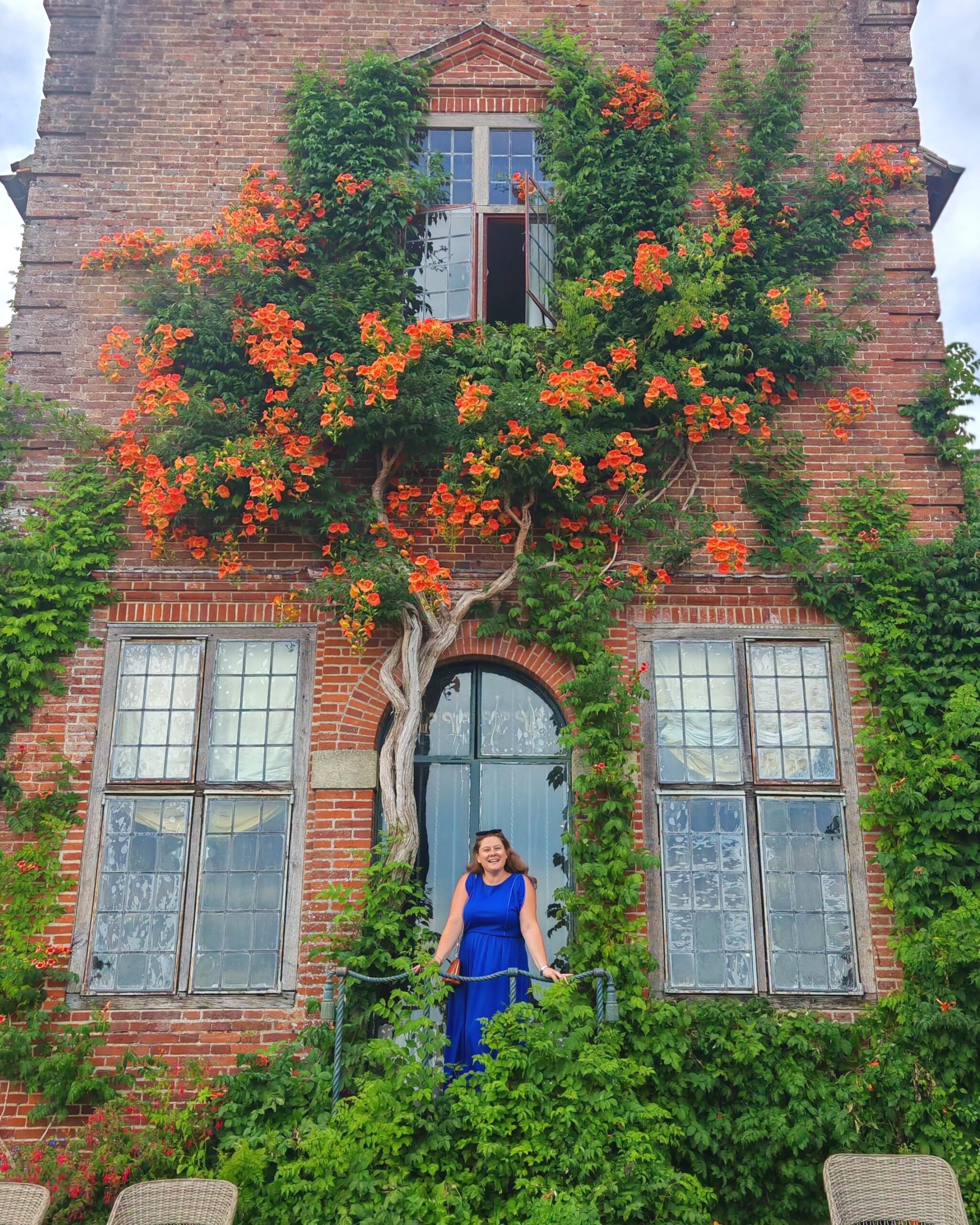 White woman in a blue dress posing by the main house at the Port Lympne Restaurant. You can see the vine with orange flowers on the wall by the balcony