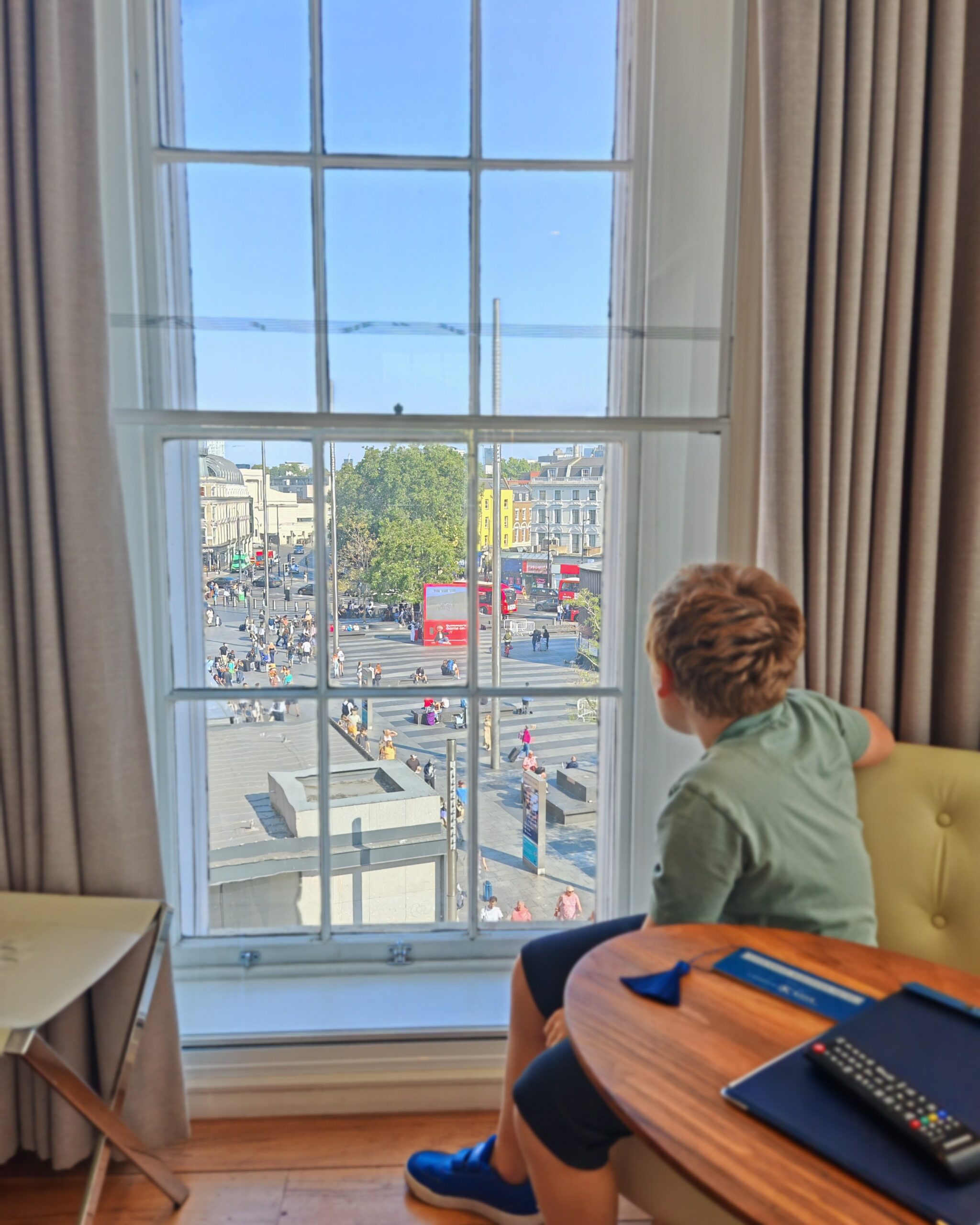 White little boy in a green tee in a room at the Kaya Great Northern Hotel in Central London. He's looking through the window, where people are catching a train at King's Cross Station.