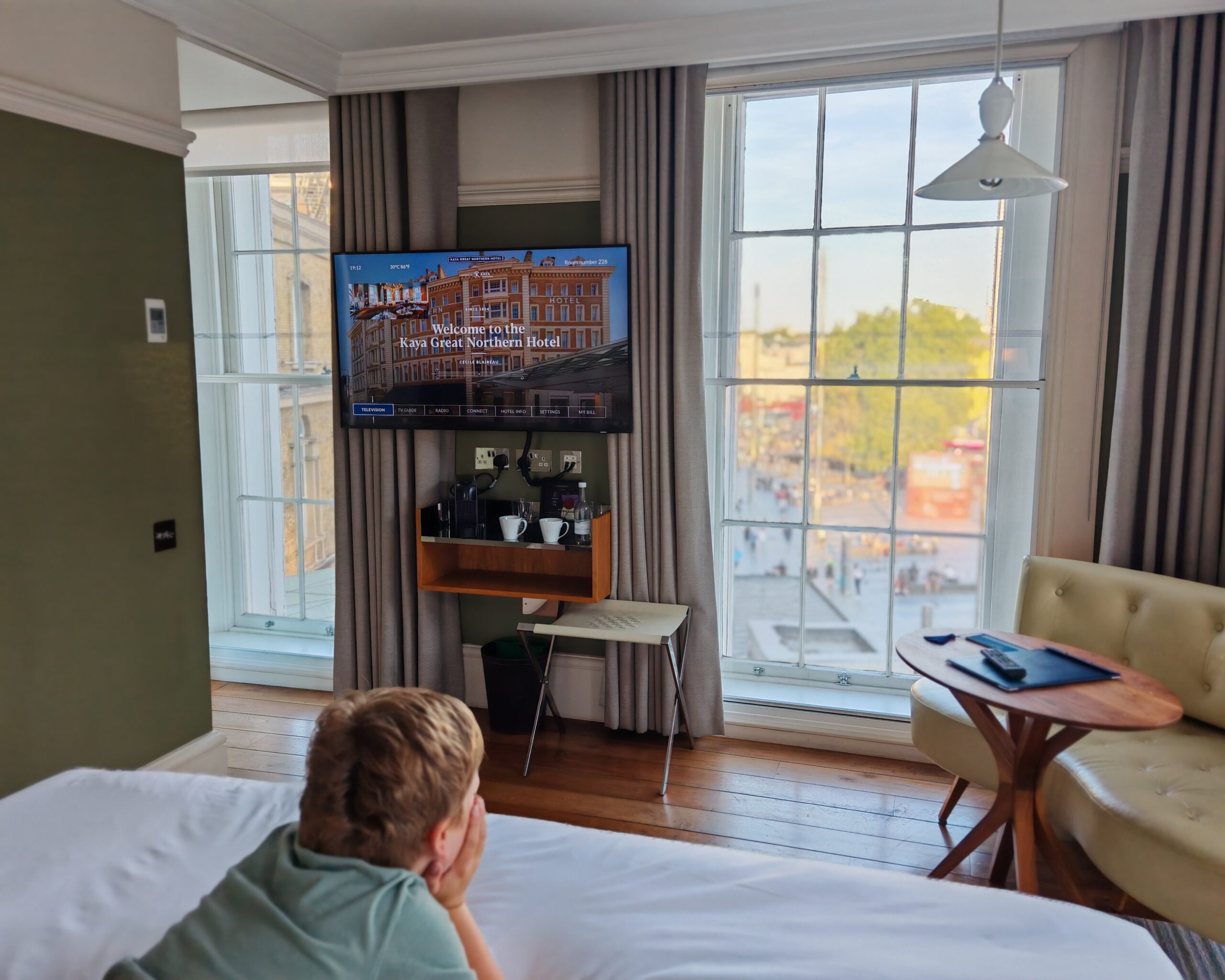 White little boy in a room at the Kaya Great Northern Hotel in Central London. He's lying on the bed, watching TV while we can see people outside the window.