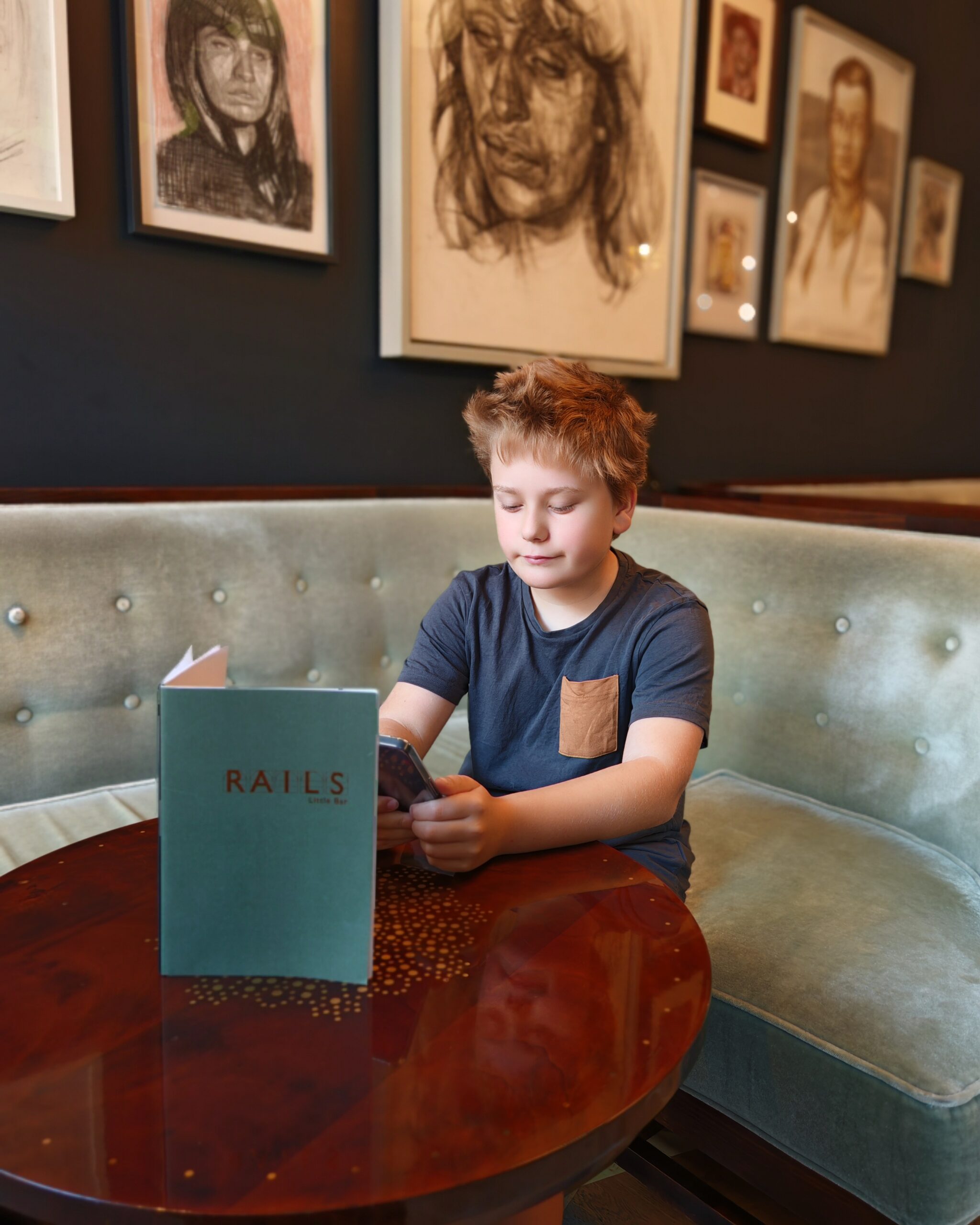 White little boy seating at the Little Bar of The Rails Restaurant in Central London. He's holding a phone right by the menu.