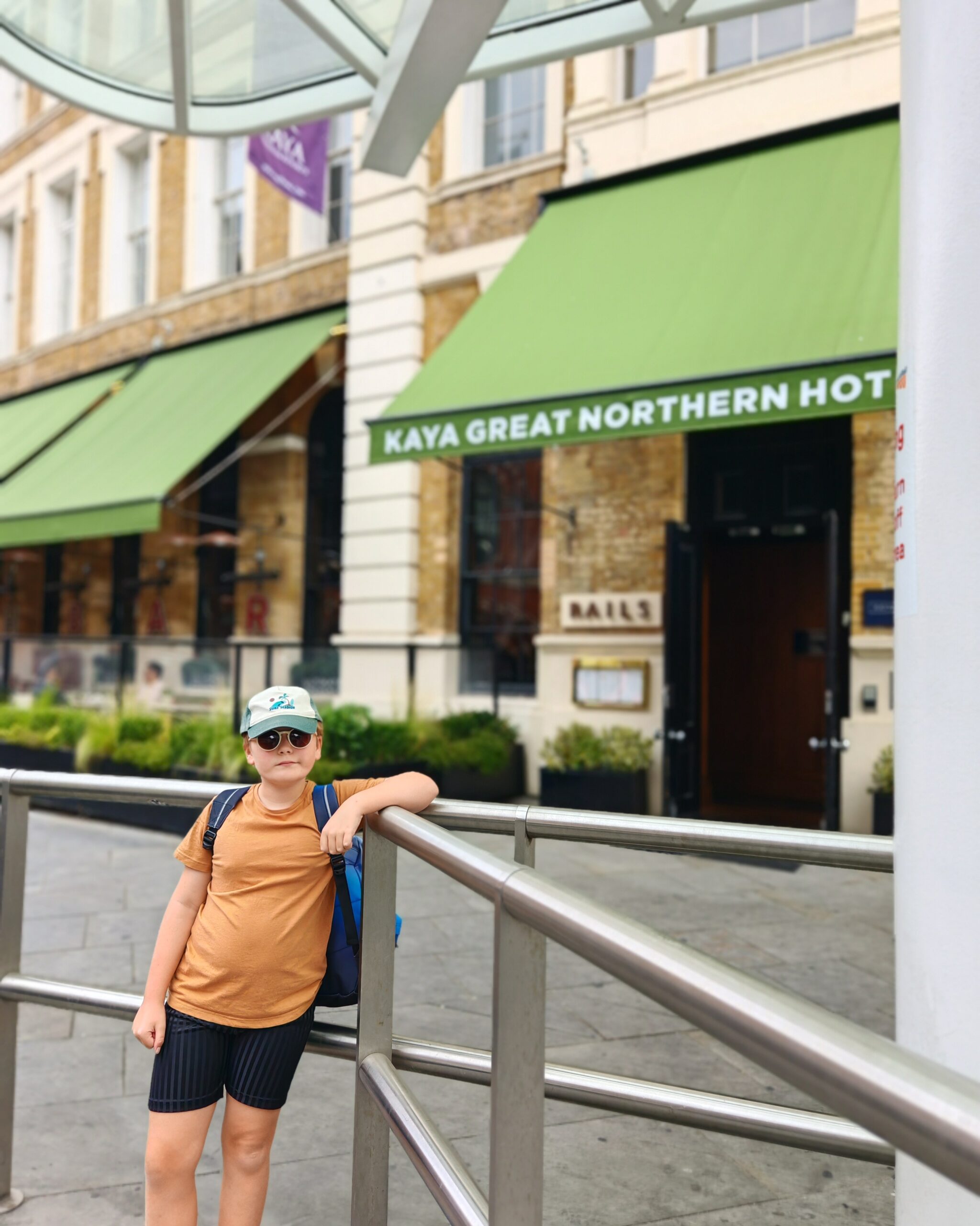 White little boy with a white cap and a yellow mustard tee standing by the taxi bay opposite the Kaya Great Northern Hotel in Central London.