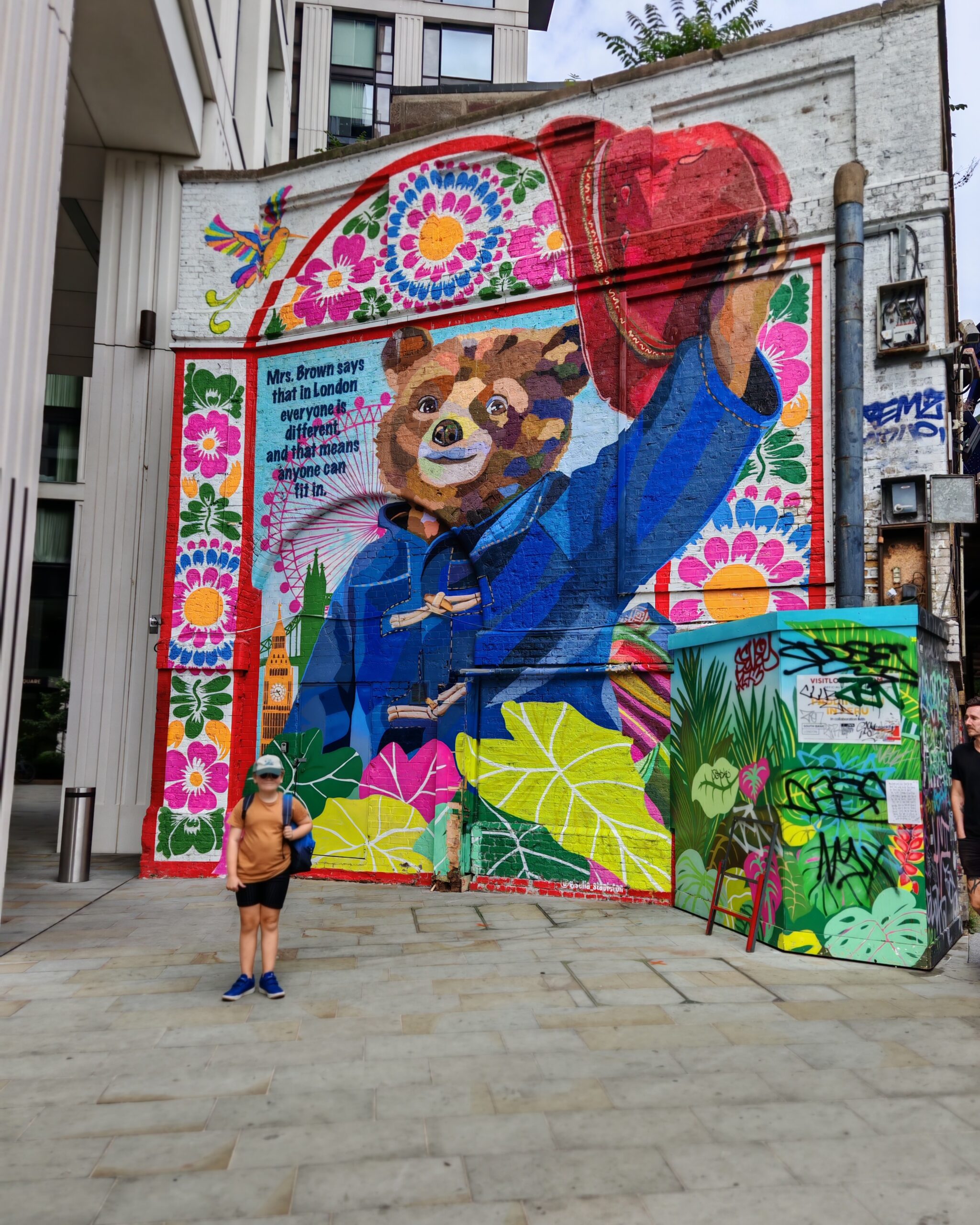 white little boy in a yellow mustard tee and black shorts posing in front of a pictural painting of Paddington on a giant wall by Waterloo Station in London