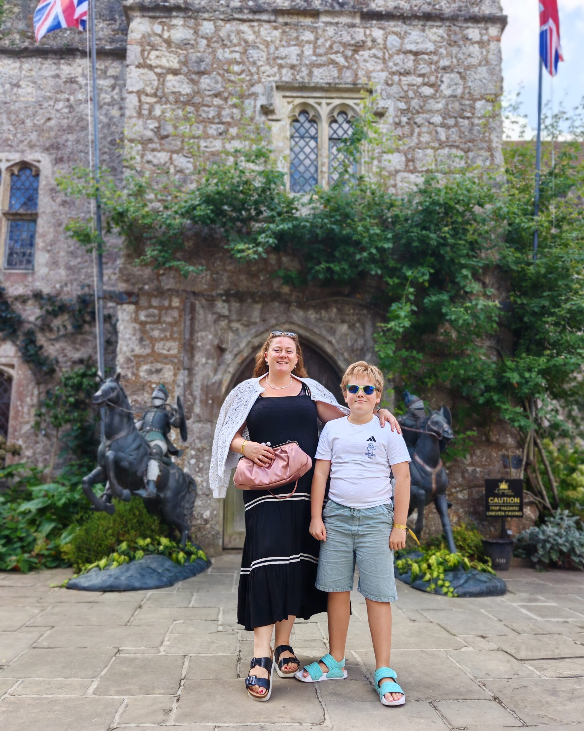 White woman in a black dress and white blazer with a little boy in white tee and green shorts in the courtyard of Lympne Castle in Kent