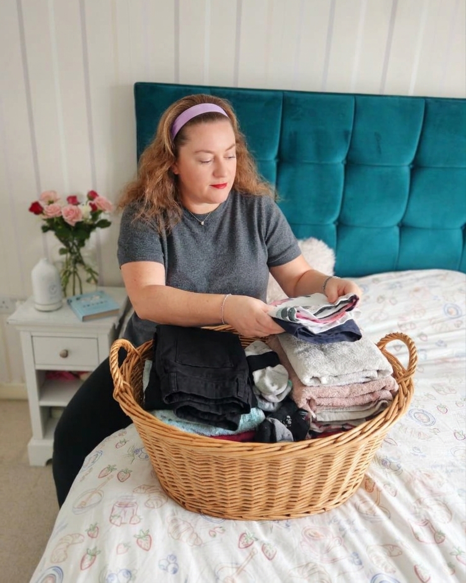 white woman with a pink hairband and a dark grey jumper sat on her bed, folding clothes with a laundry basket next to her